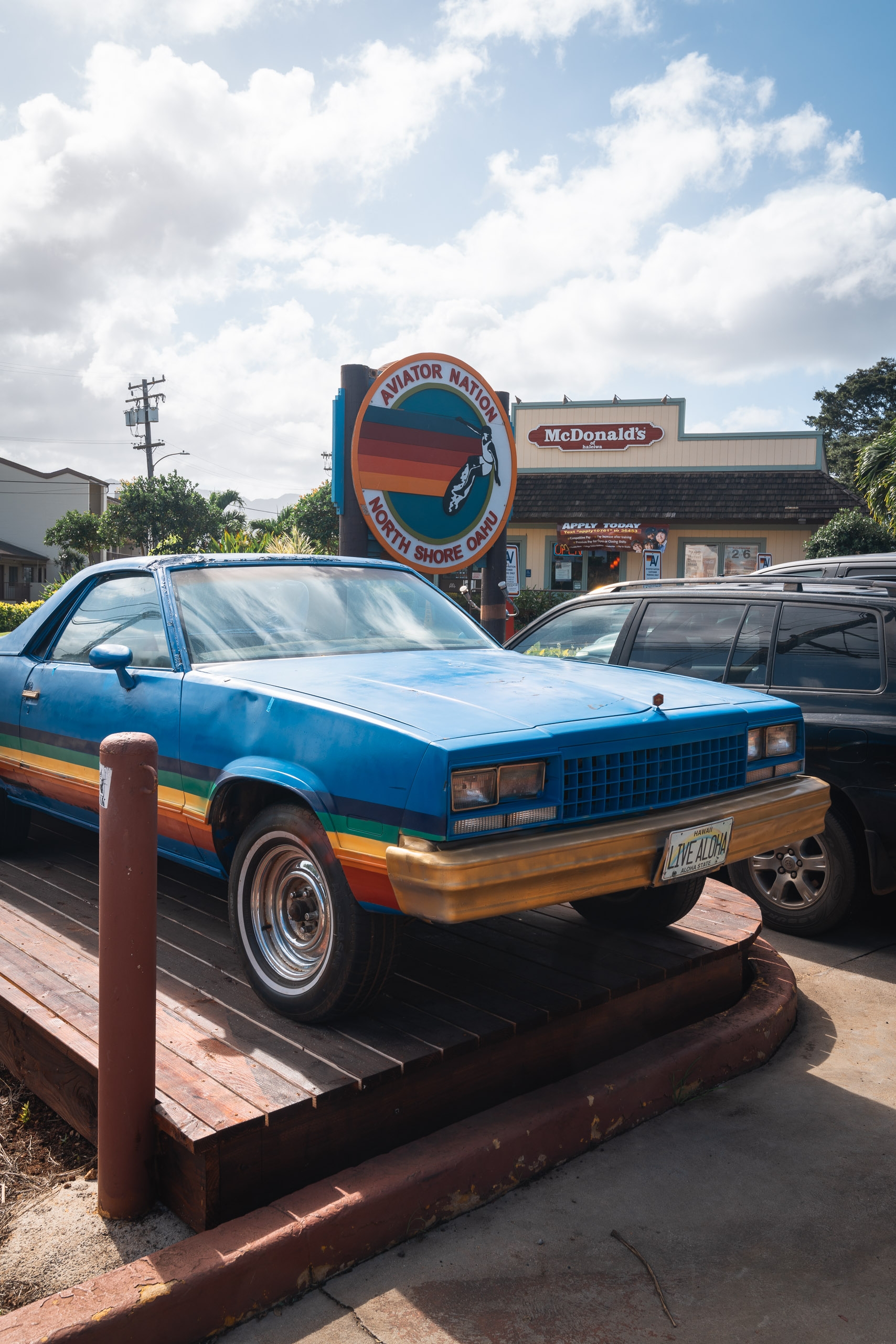 A blue and wood-paneled vintage car sits parked outside an Aviator Nation surf shop near a McDonald's, under bright Hawaiian skies with white clouds