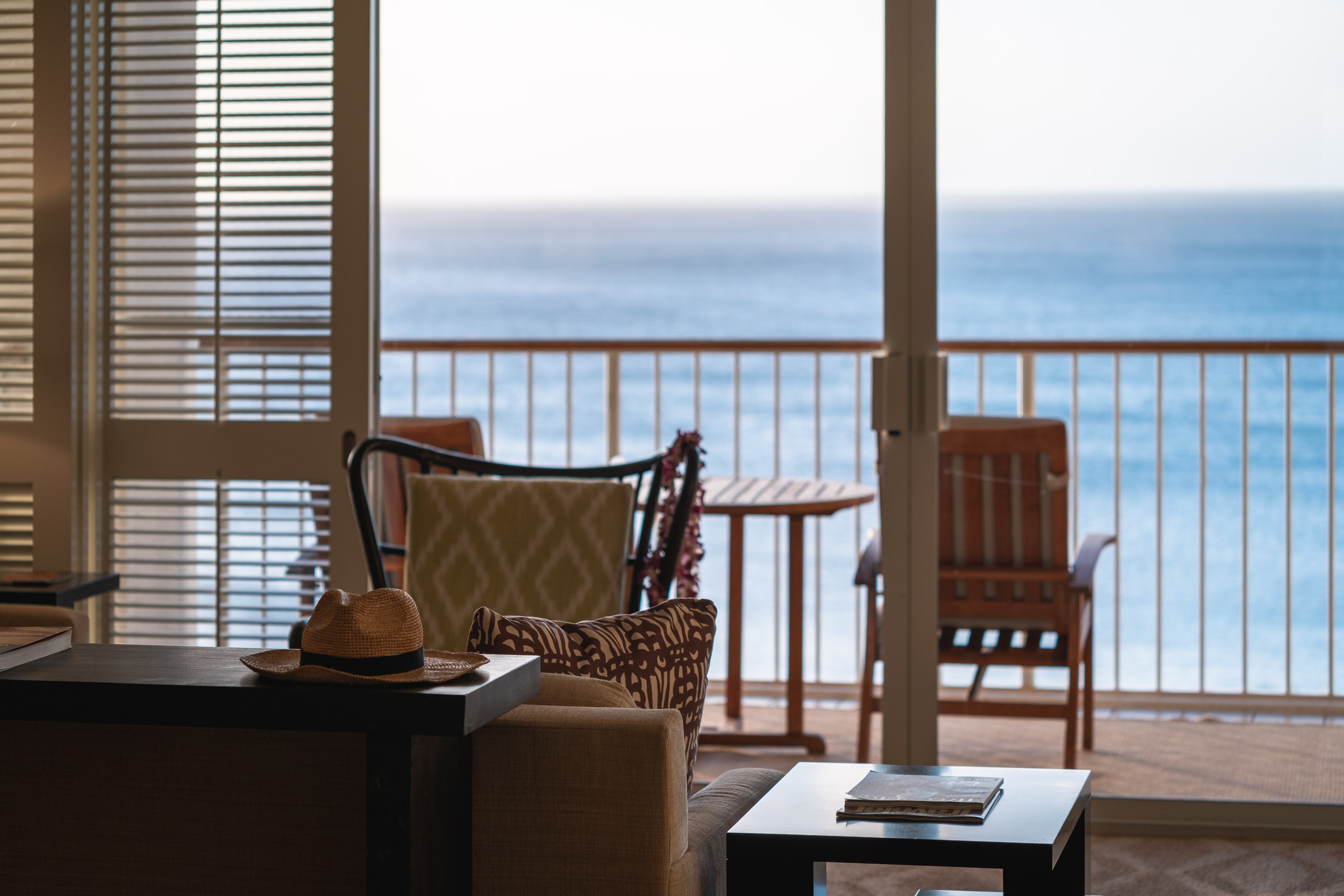 A luxury beachfront suite features comfortable seating, a small table with a straw hat, and expansive ocean views framed by glass doors and metal railings under soft daylight