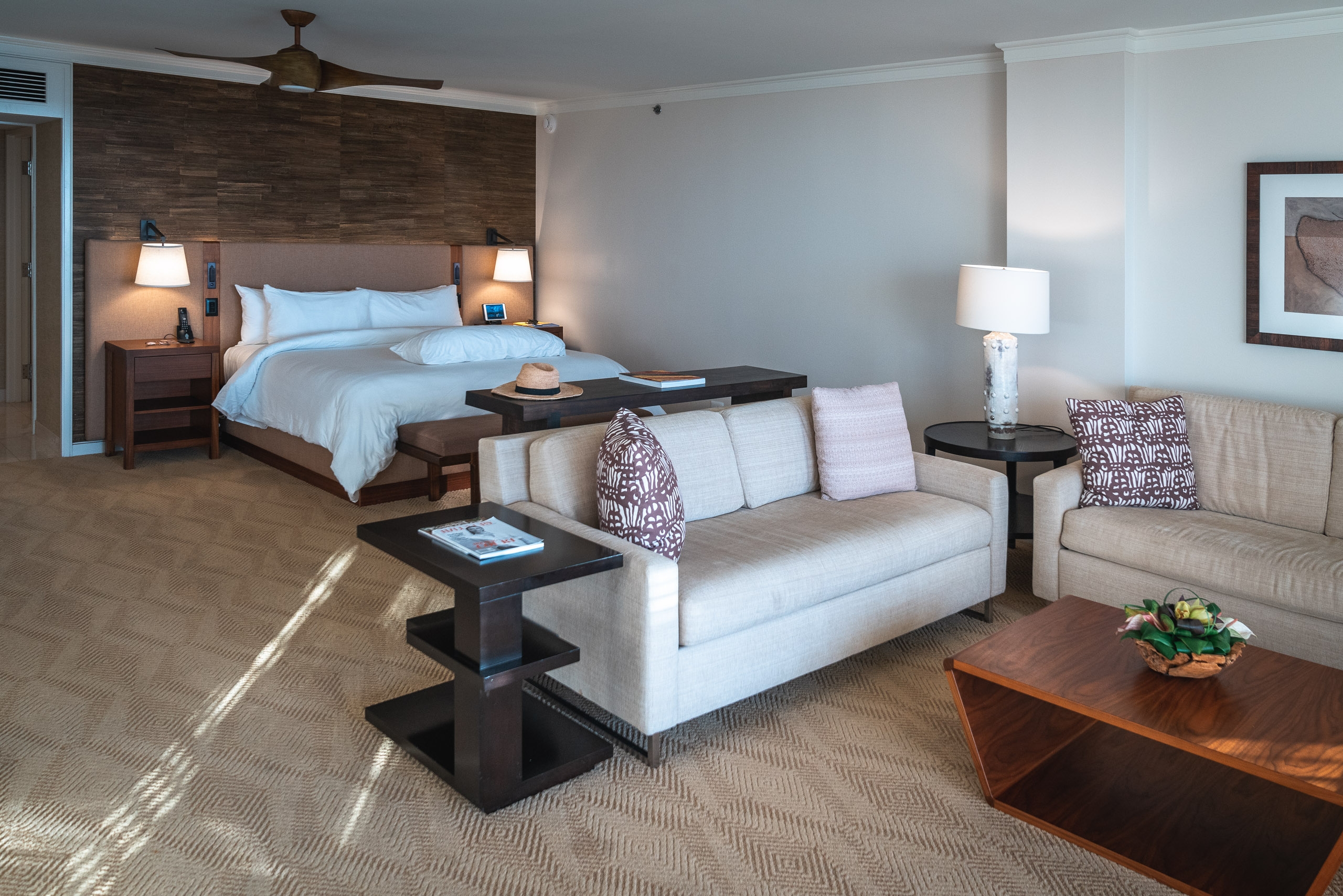 A spacious hotel suite features a king bed with blue linens and wood accent wall, a cream-colored sofa with patterned pillows, and warm lighting from table lamps creating an inviting contemporary resort interior