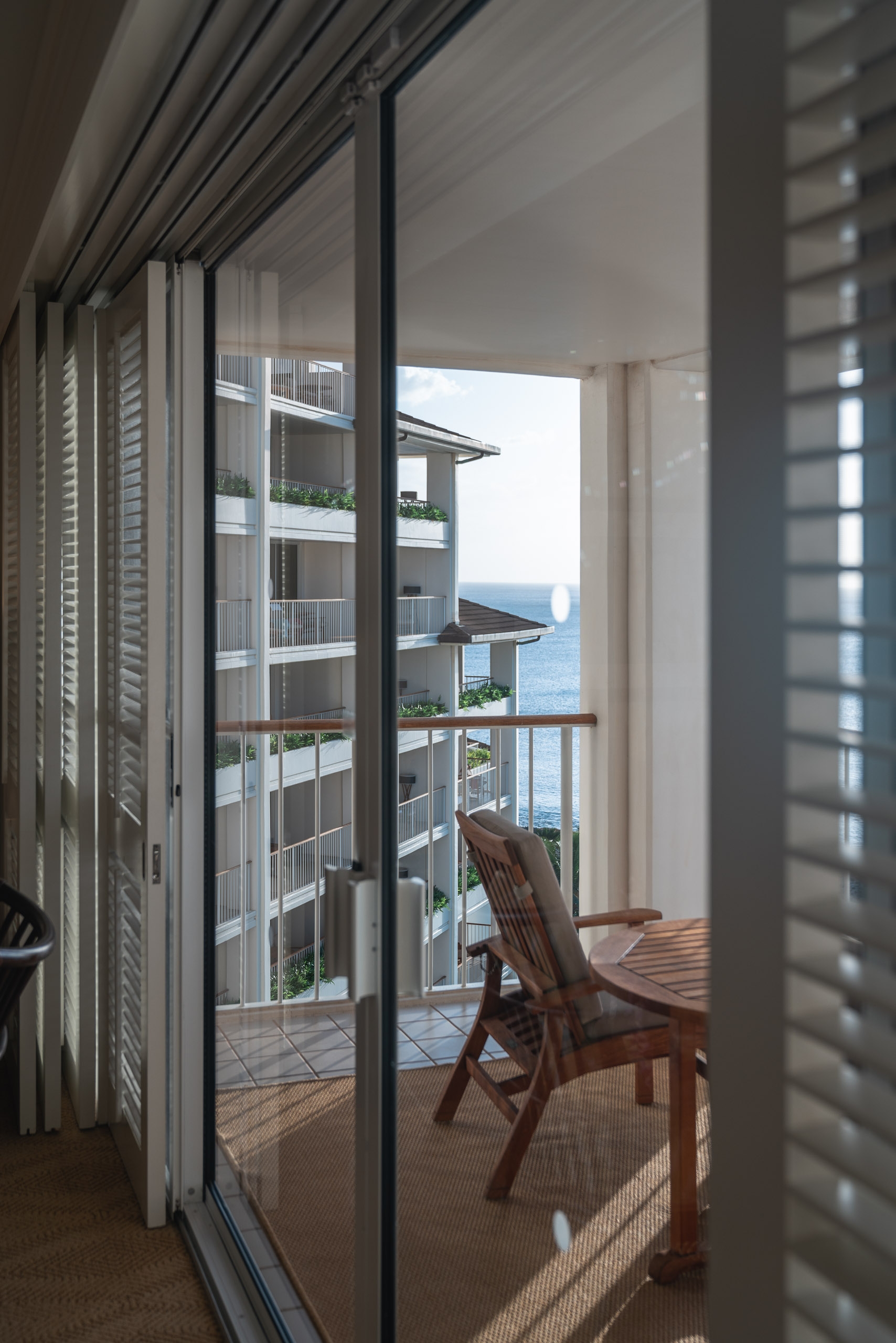 Wooden lounge chair sits on a sunlit oceanfront balcony framed by sliding glass doors and white louvered shutters, with distant mountains visible across the water