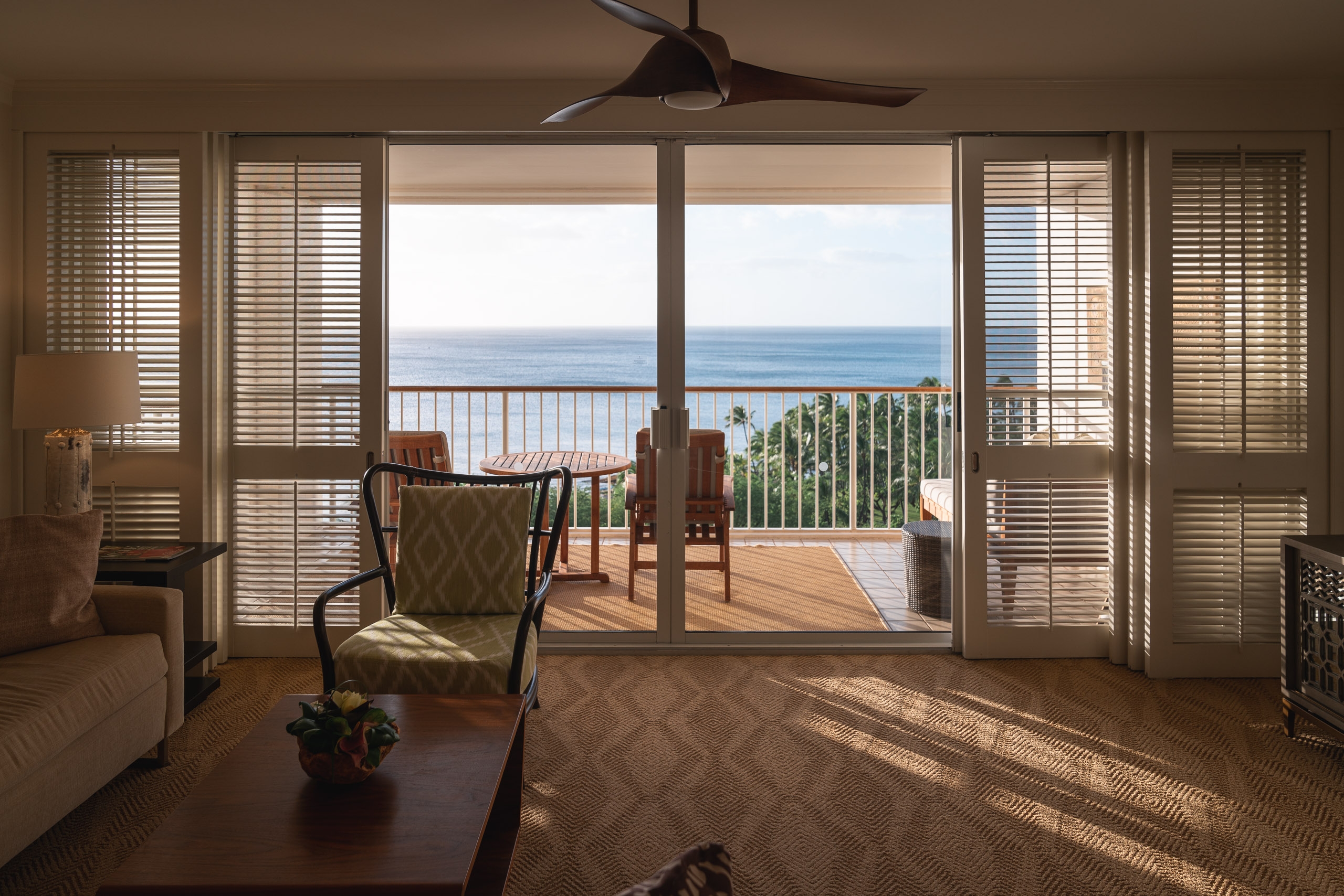 Bright sunlight streams through expansive glass doors illuminating a luxury oceanfront hotel suite with wooden shutters, comfortable seating, and a panoramic view of the blue Pacific Ocean beyond the railinged balcony
