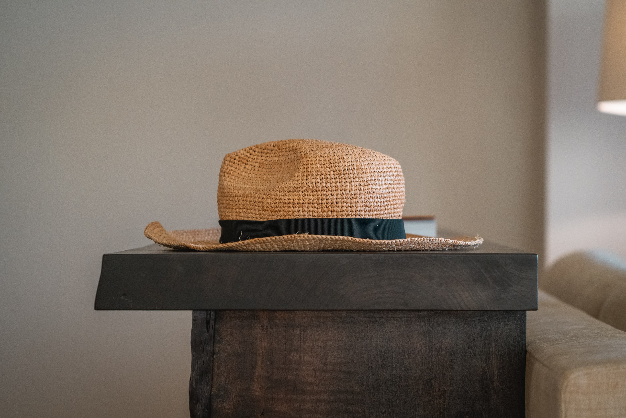 A woven straw hat with a dark band sits atop a dark wooden pedestal in soft, diffused indoor light with neutral walls