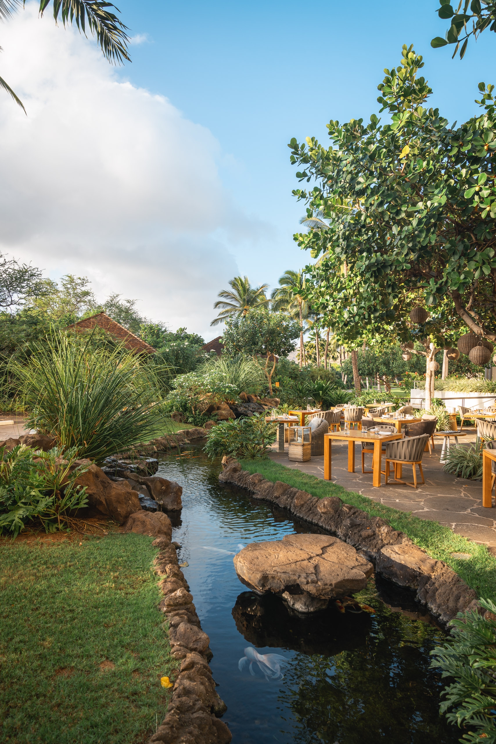 A tranquil resort garden features a small stream with rocky banks winding past manicured lawns and tropical vegetation toward wooden dining tables beneath palm and shade trees under clear blue sky
