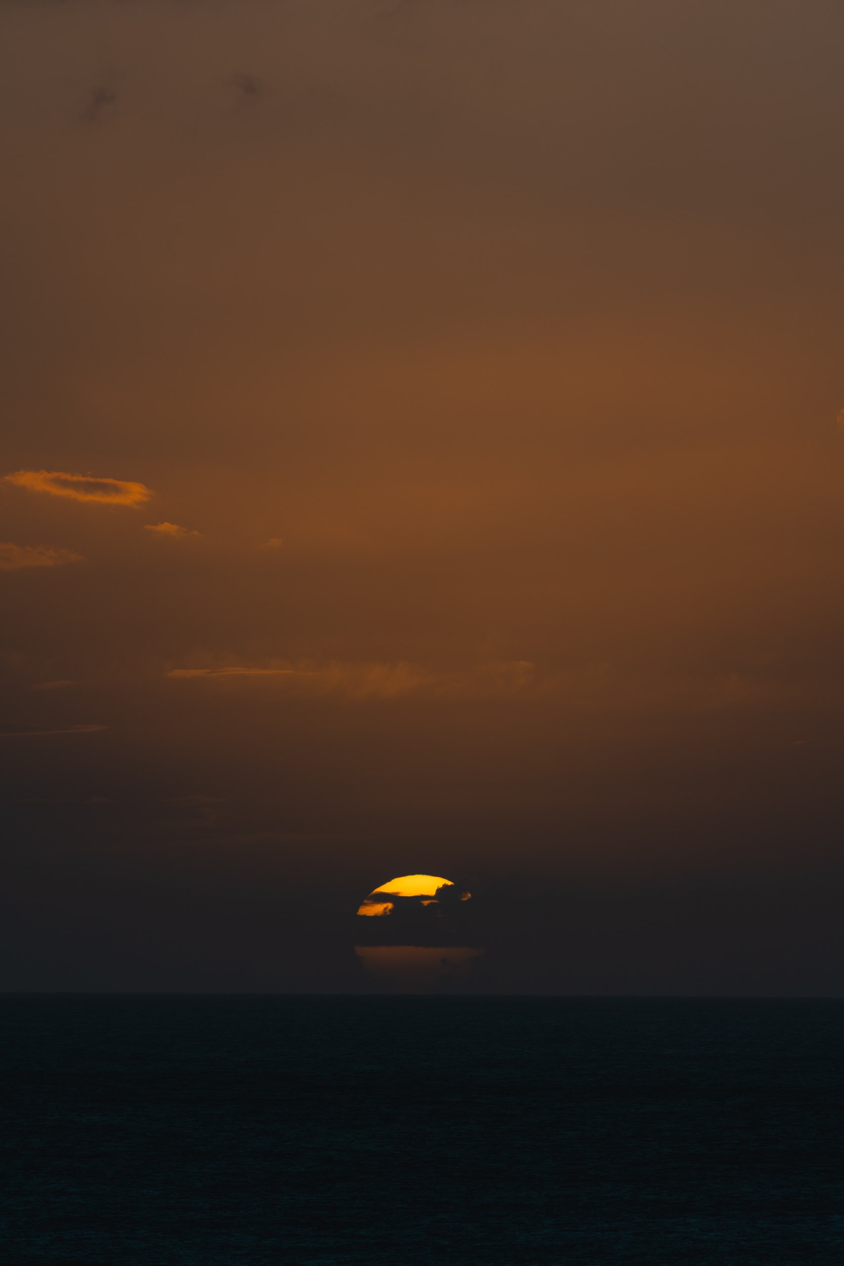 A golden sun descends toward the dark ocean horizon at dusk, casting warm amber tones across a layered sky that transitions from brown to deep blue-black
