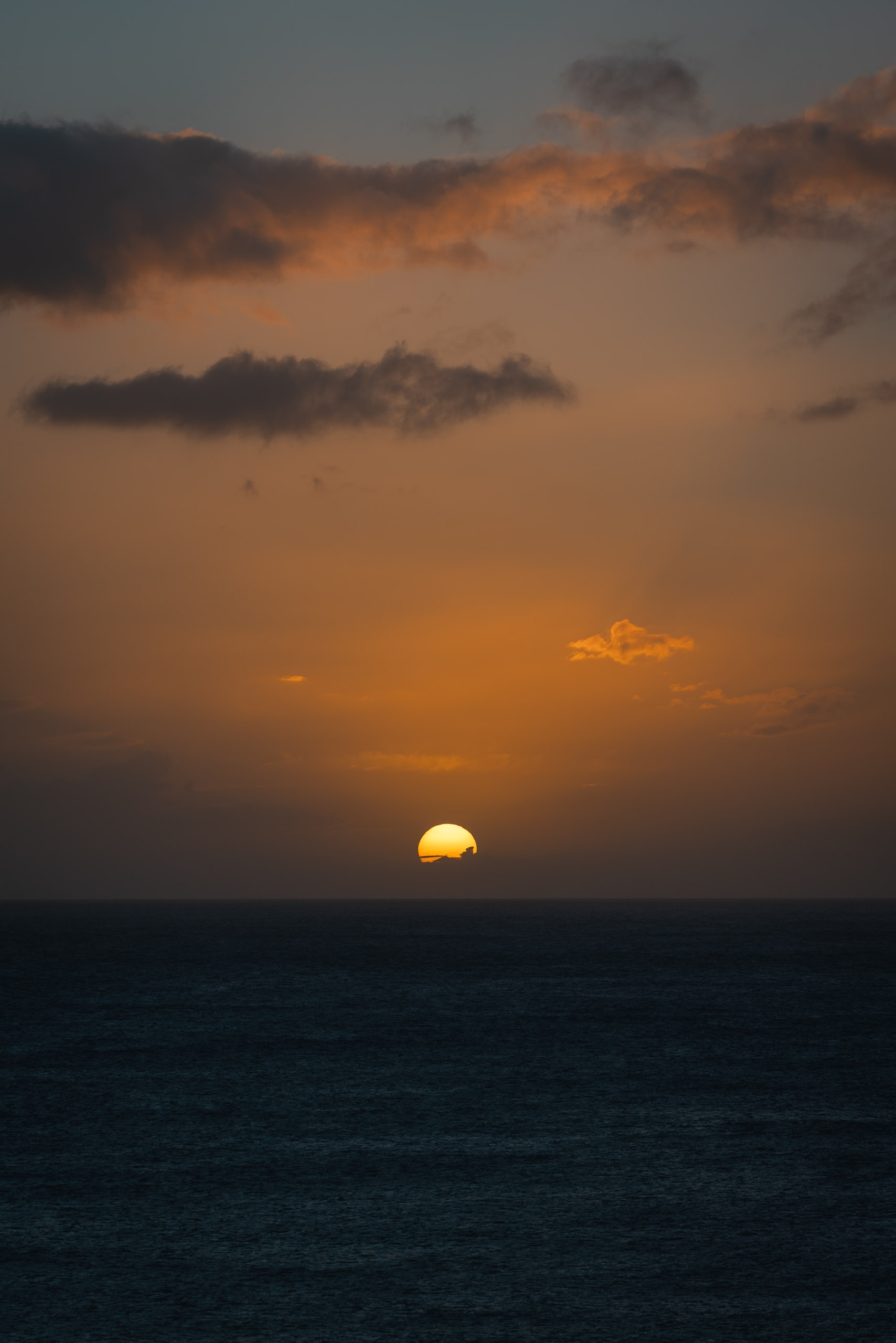 Golden sun sinks toward the dark ocean horizon beneath layered clouds in warm orange and amber tones at dusk