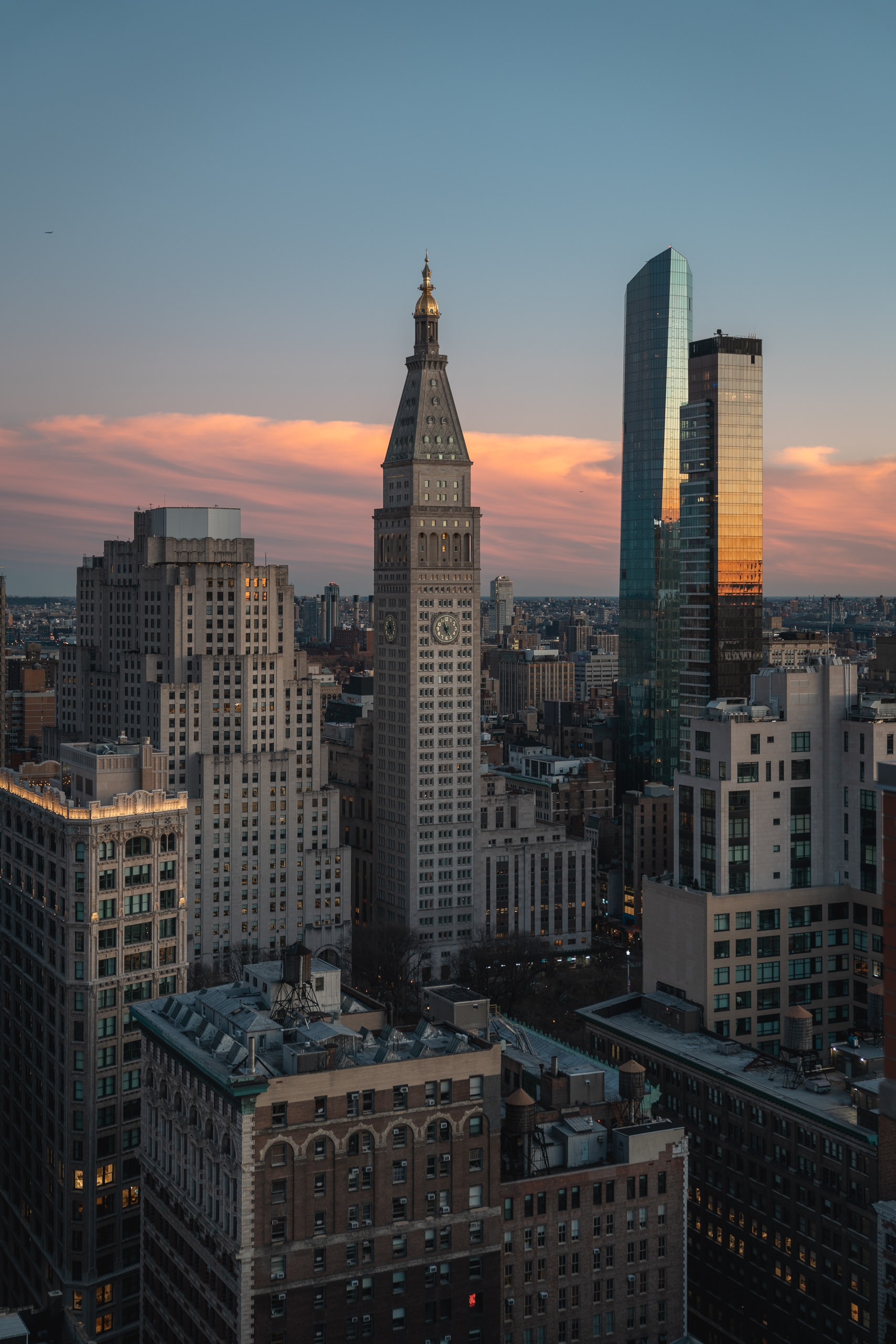 Manhattan skyline at dusk features art deco towers with golden spire in center, modern glass skyscraper on right, warm sunset light illuminating building facades and clouds above