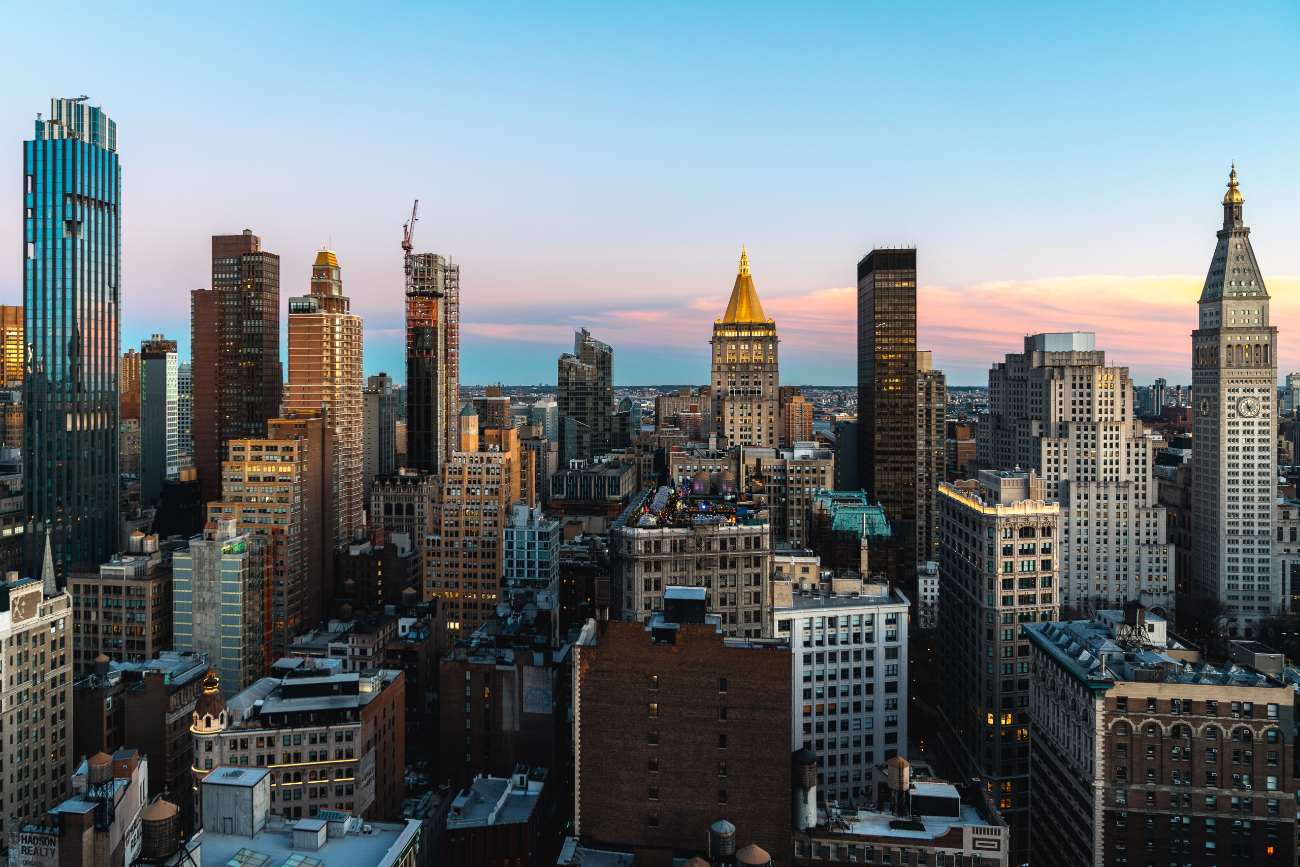 Manhattan's densely packed skyline stretches across the frame under a twilight sky painted in soft pink and orange hues, with iconic Art Deco and modern skyscrapers catching golden light