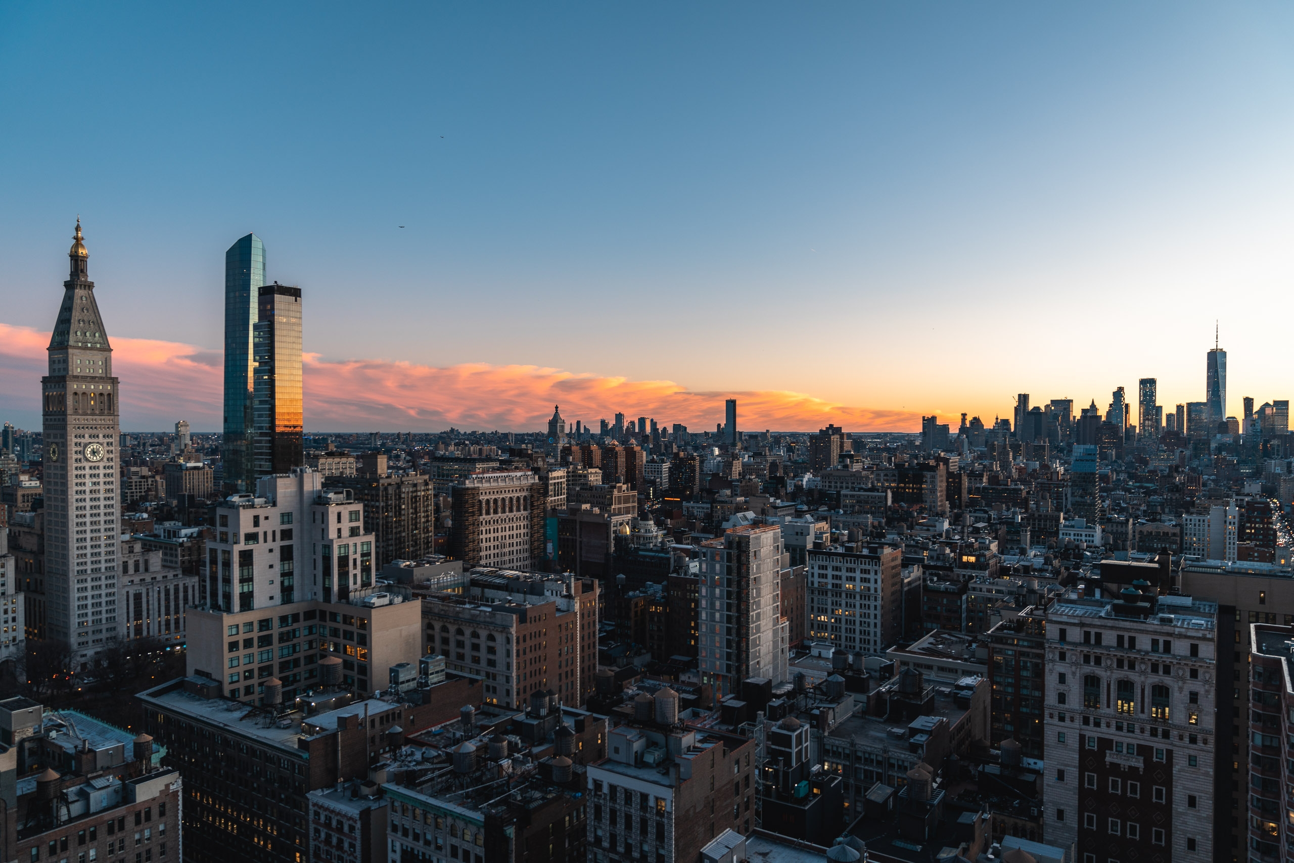 Manhattan's dense skyline stretches toward the horizon under an orange and blue sunset, with the Chrysler Building prominent on the left and modern skyscrapers glowing against darkening clouds