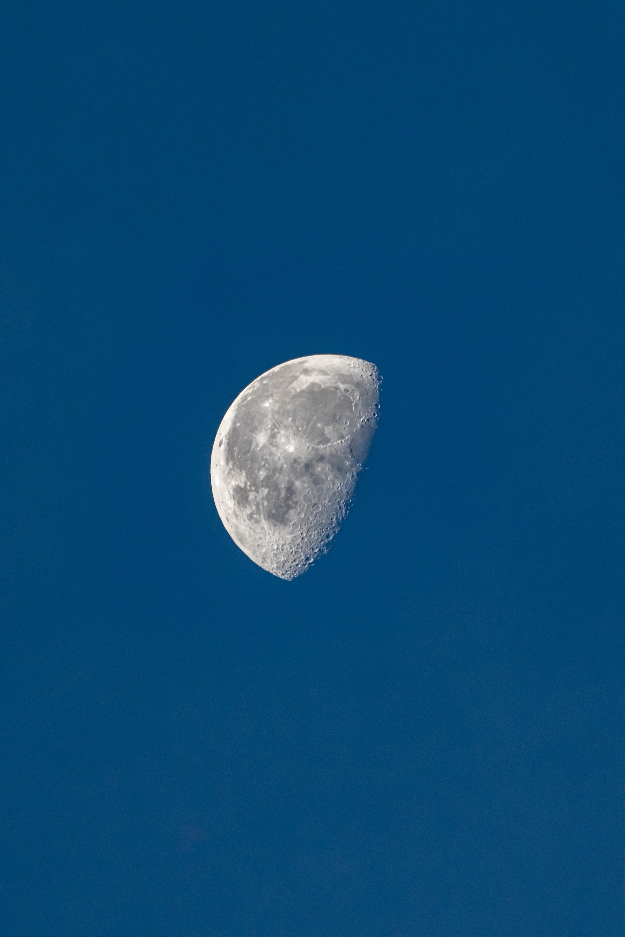 A waning crescent moon with visible surface craters and detail hangs against a deep blue daytime sky, photographed from Earth during daylight hours