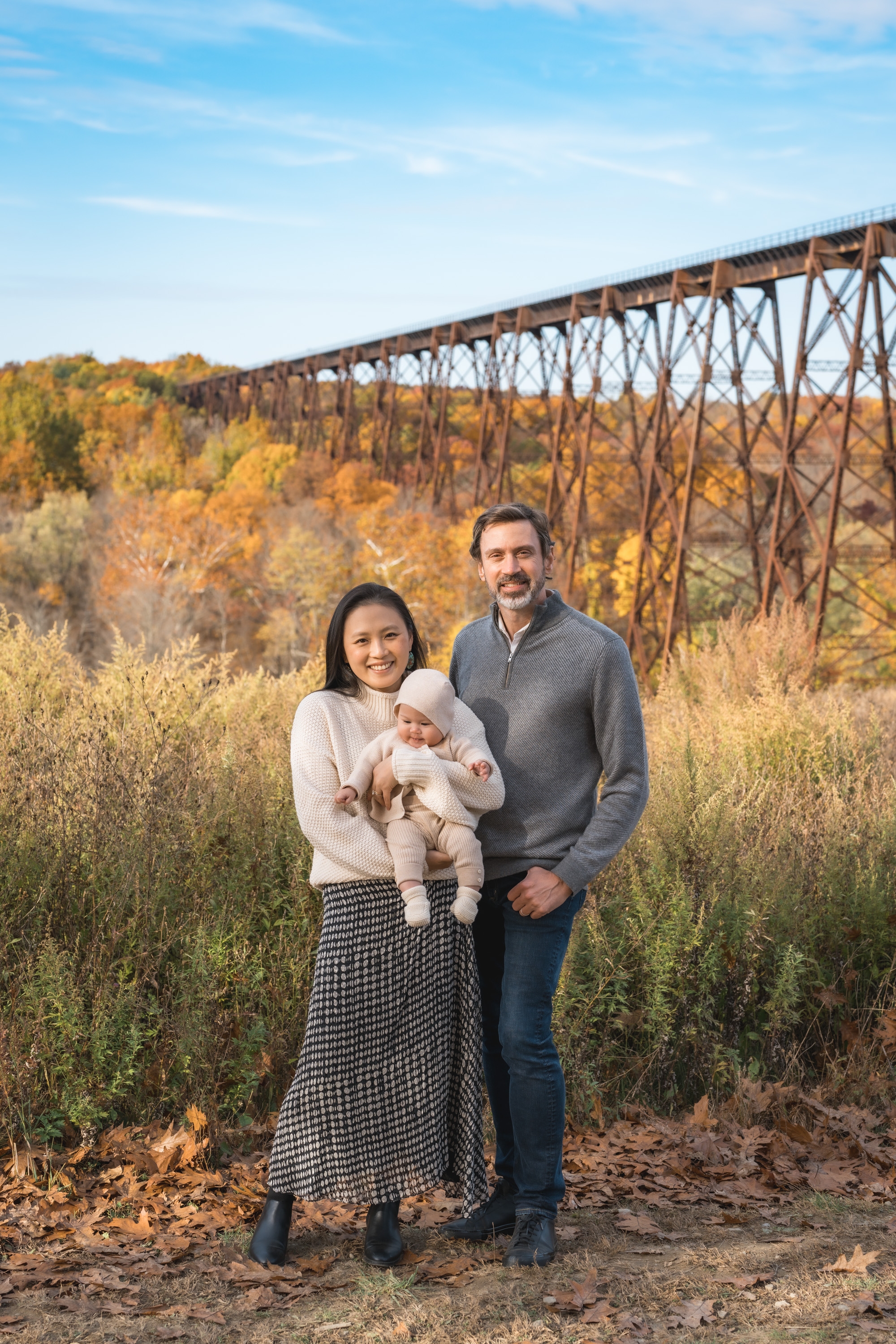 A family of three stands in an autumn landscape with golden foliage and bare trees, with a tall iron railroad trestle bridge stretching across the valley behind them under a clear blue sky