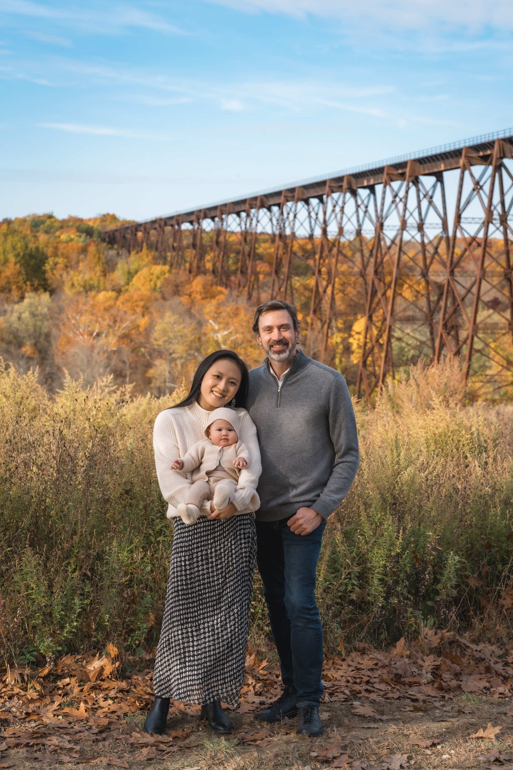 A family of three stands in an autumn field with golden foliage and a tall trestle bridge spanning the valley behind them under clear blue sky