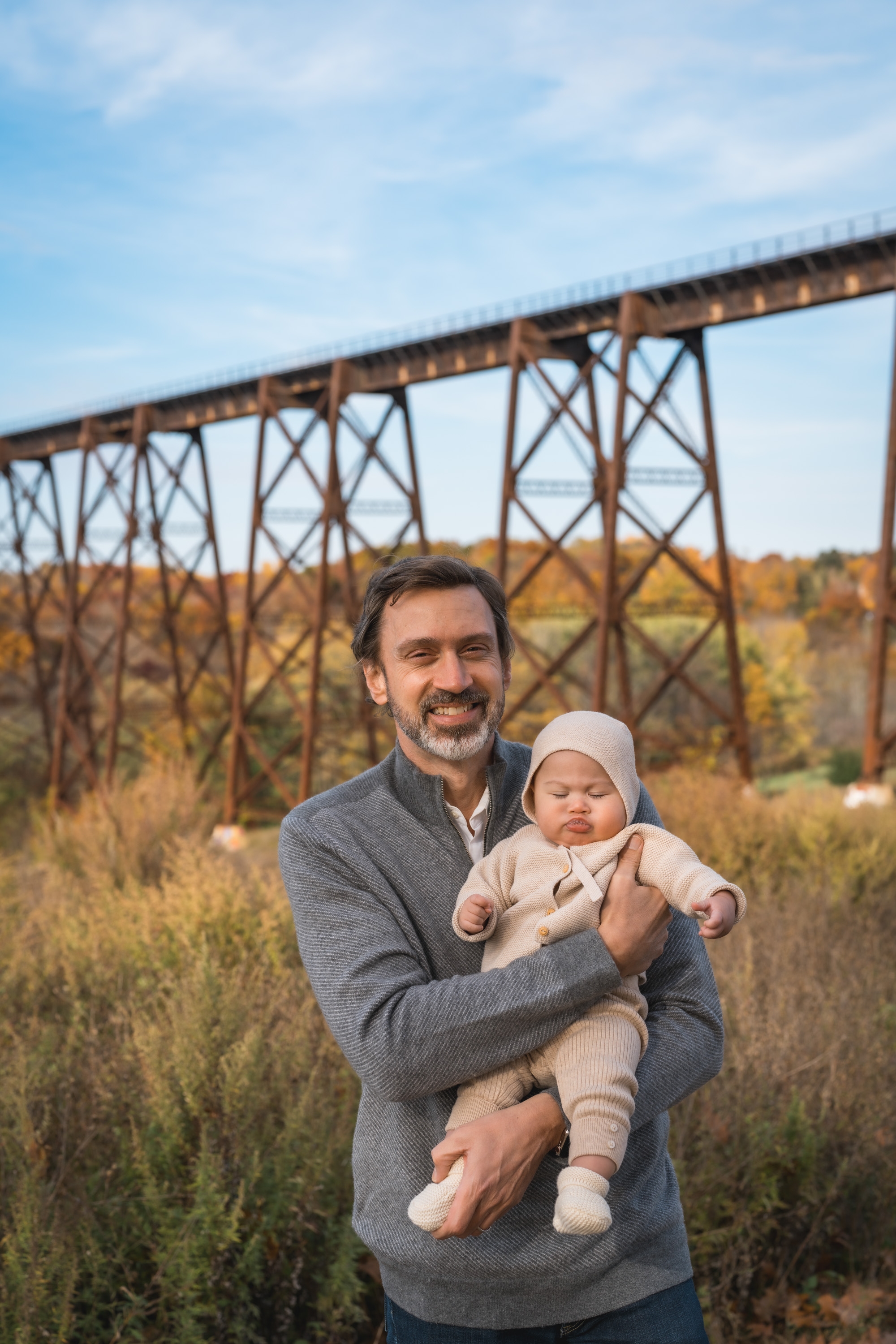 A man holds his infant daughter while standing in golden autumn vegetation, with a towering rusty railroad trestle bridge stretching across the background beneath a partly cloudy sky