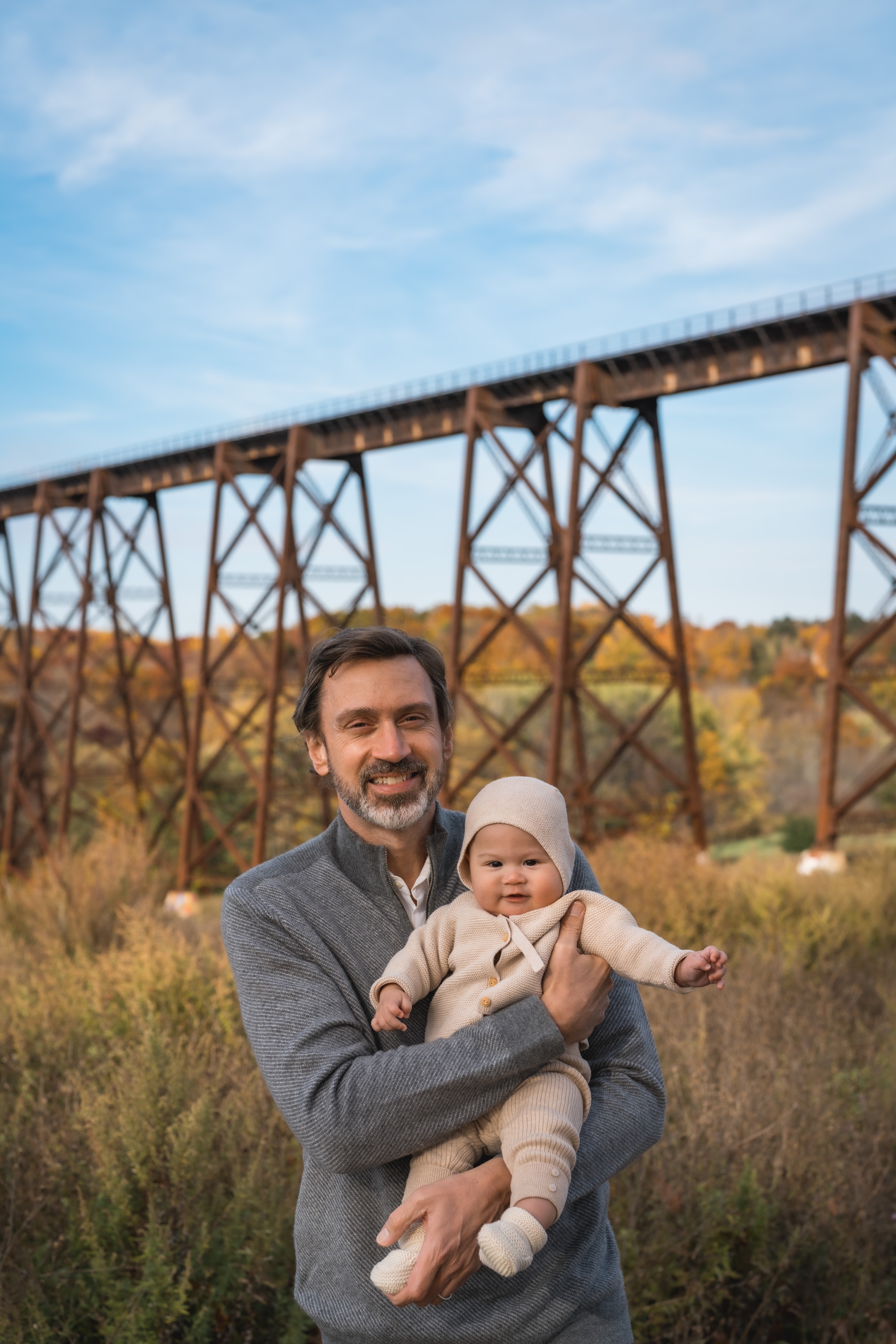 A bearded man in a gray cardigan holds a smiling infant wearing a white hat and cream-colored outfit, with a towering rusted steel trestle bridge and autumn foliage visible behind them under a partly cloudy sky