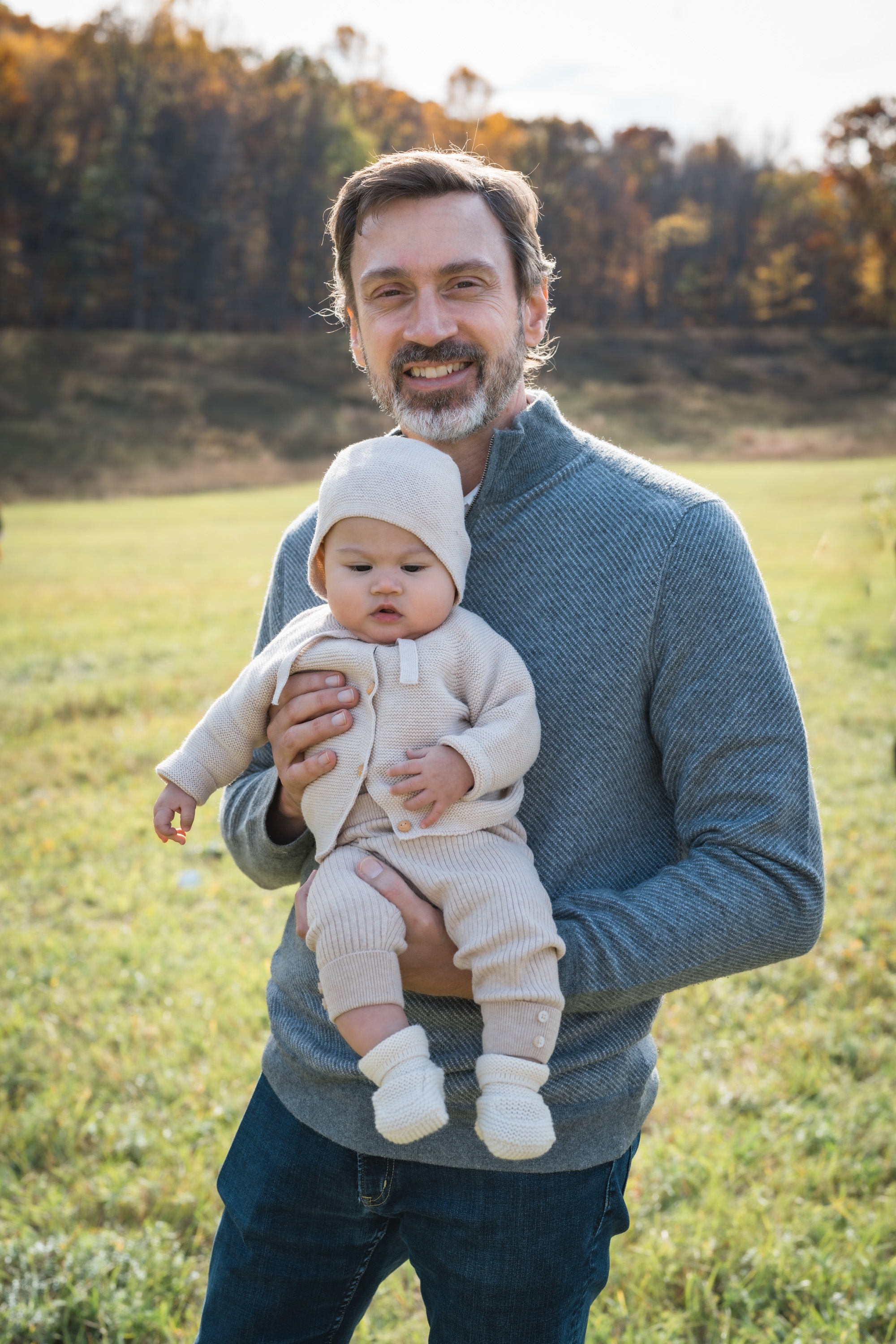 A man holding an infant in cream-colored clothing stands in an open field with autumn-toned forest and golden light behind them.