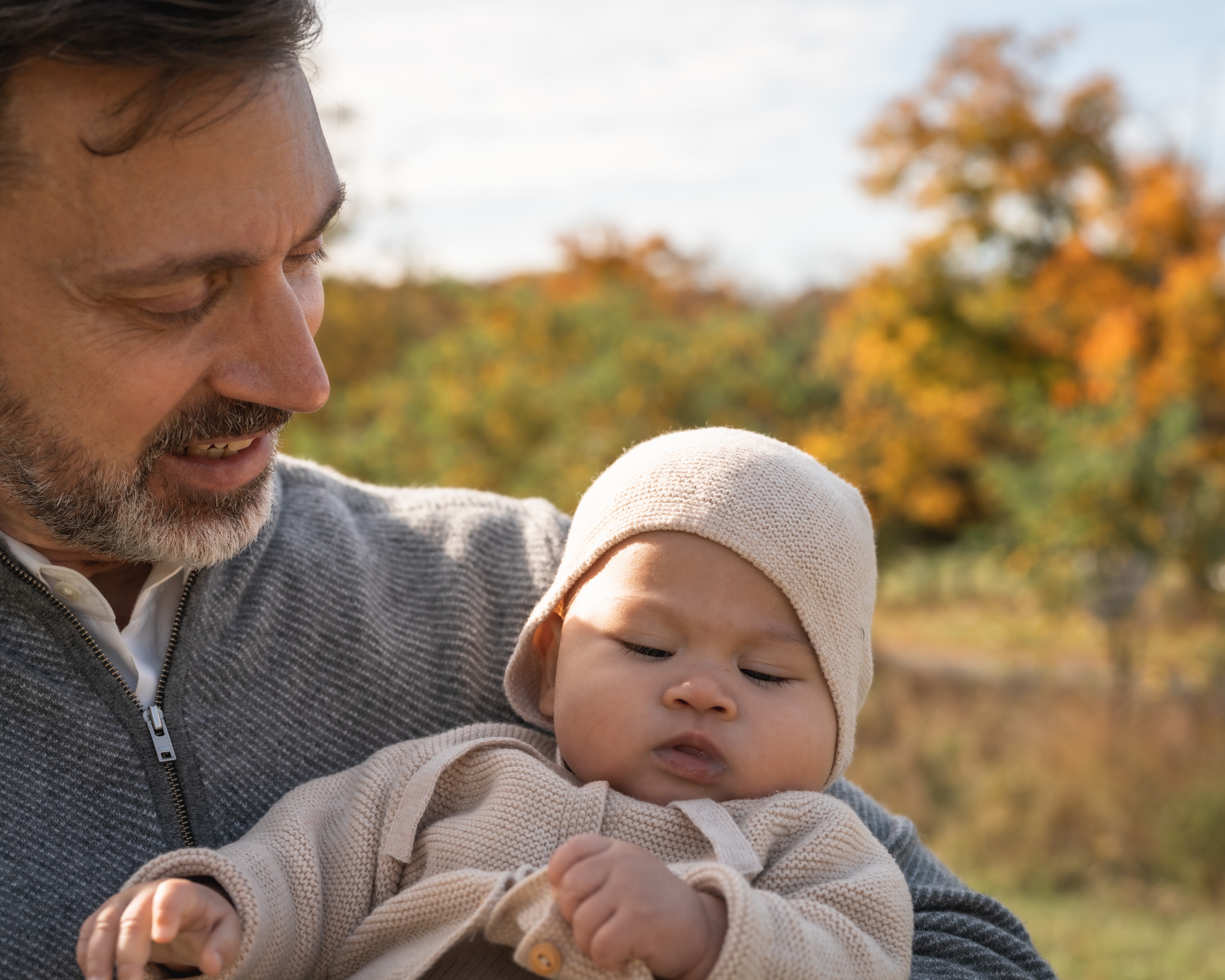 A bearded man holds a sleeping infant in a cream knit hat and sweater against an autumn landscape with golden foliage and overcast sky