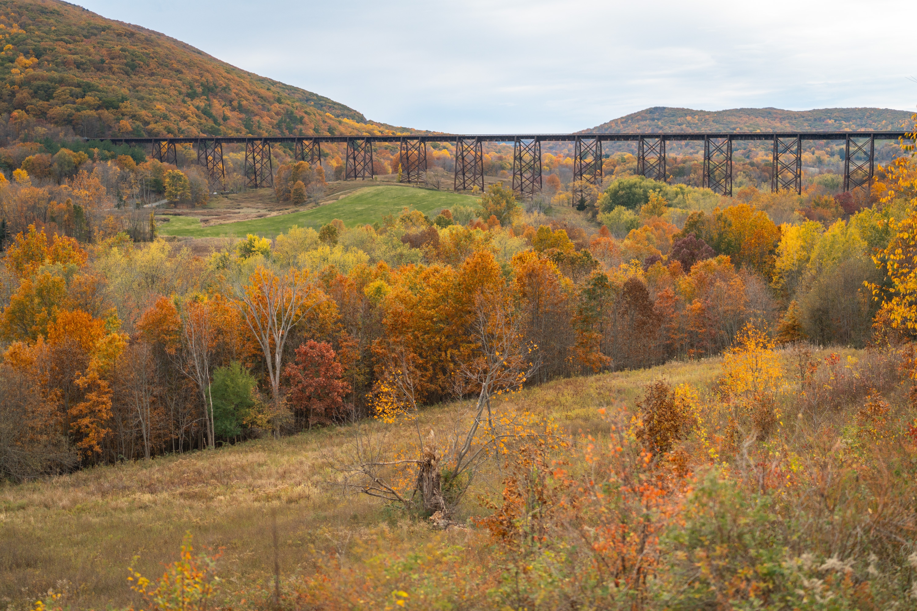 A tall iron railroad trestle spans a forested valley during autumn, with golden and orange deciduous trees covering rolling hills beneath an overcast sky