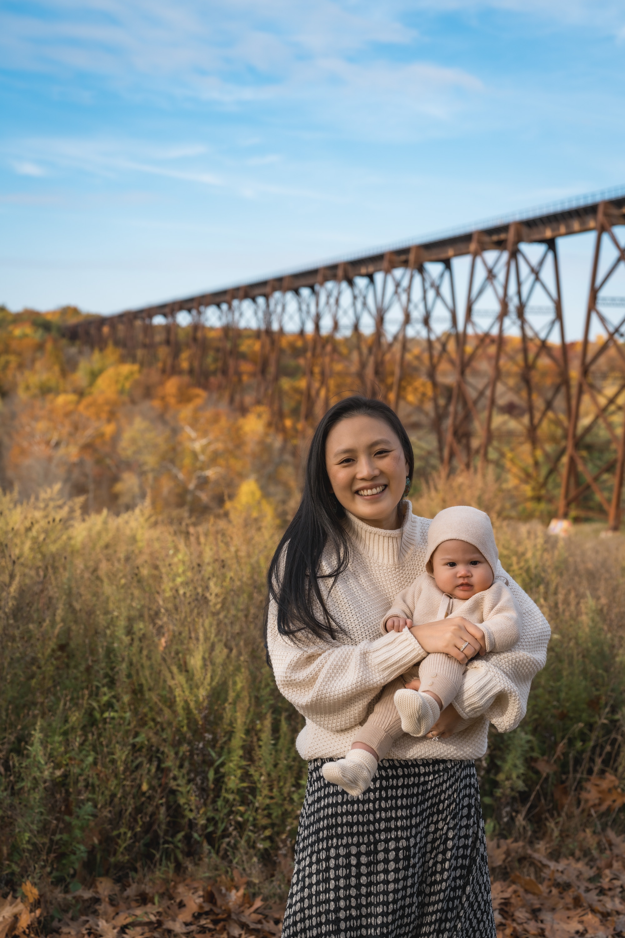 A woman holding an infant in cream-colored knitwear stands before an iron trestle bridge with autumn foliage below under clear blue sky