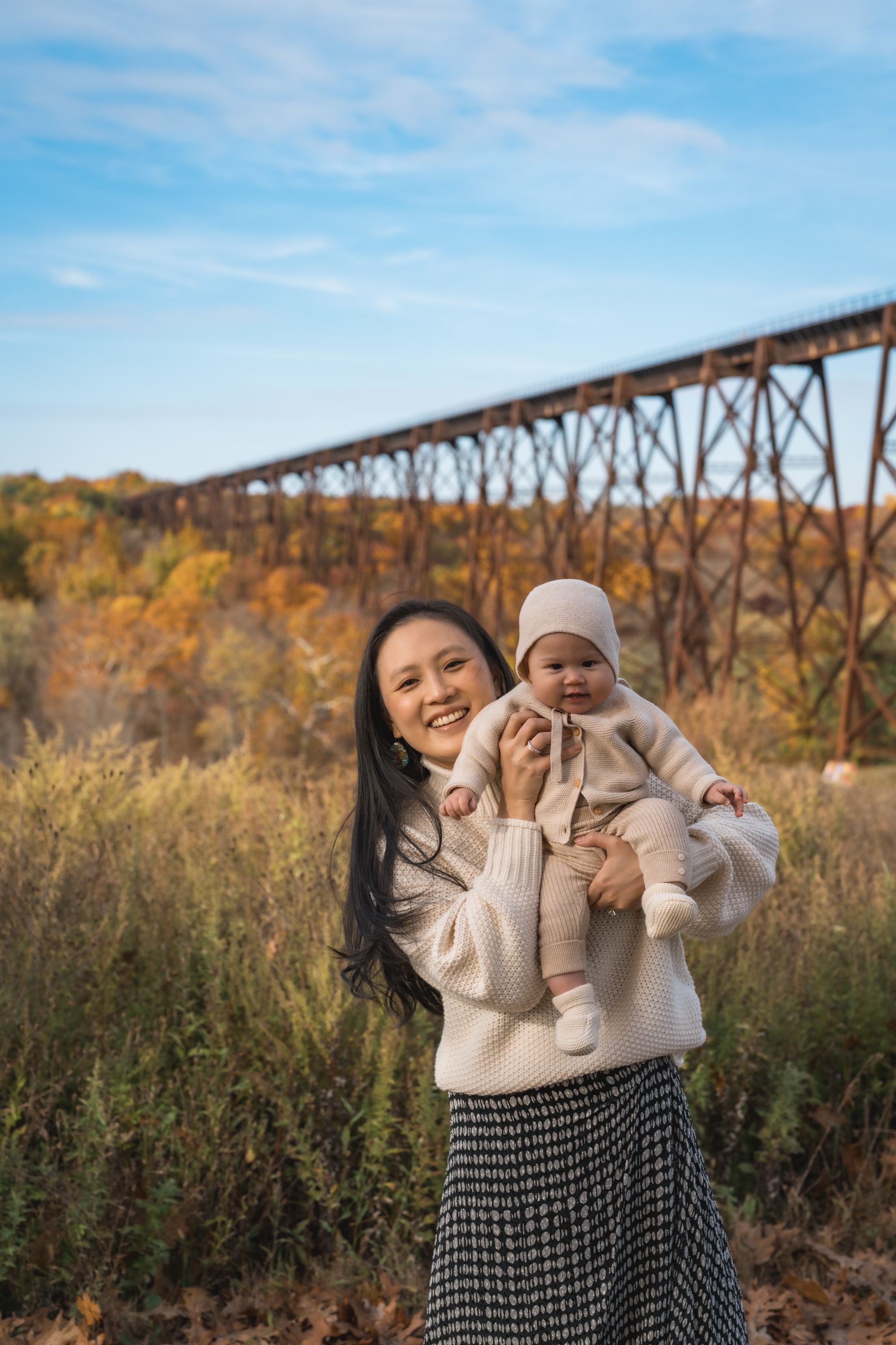 Woman holding an infant in cream-colored clothing stands before a rustic iron trestle bridge, surrounded by golden autumn foliage under clear blue sky