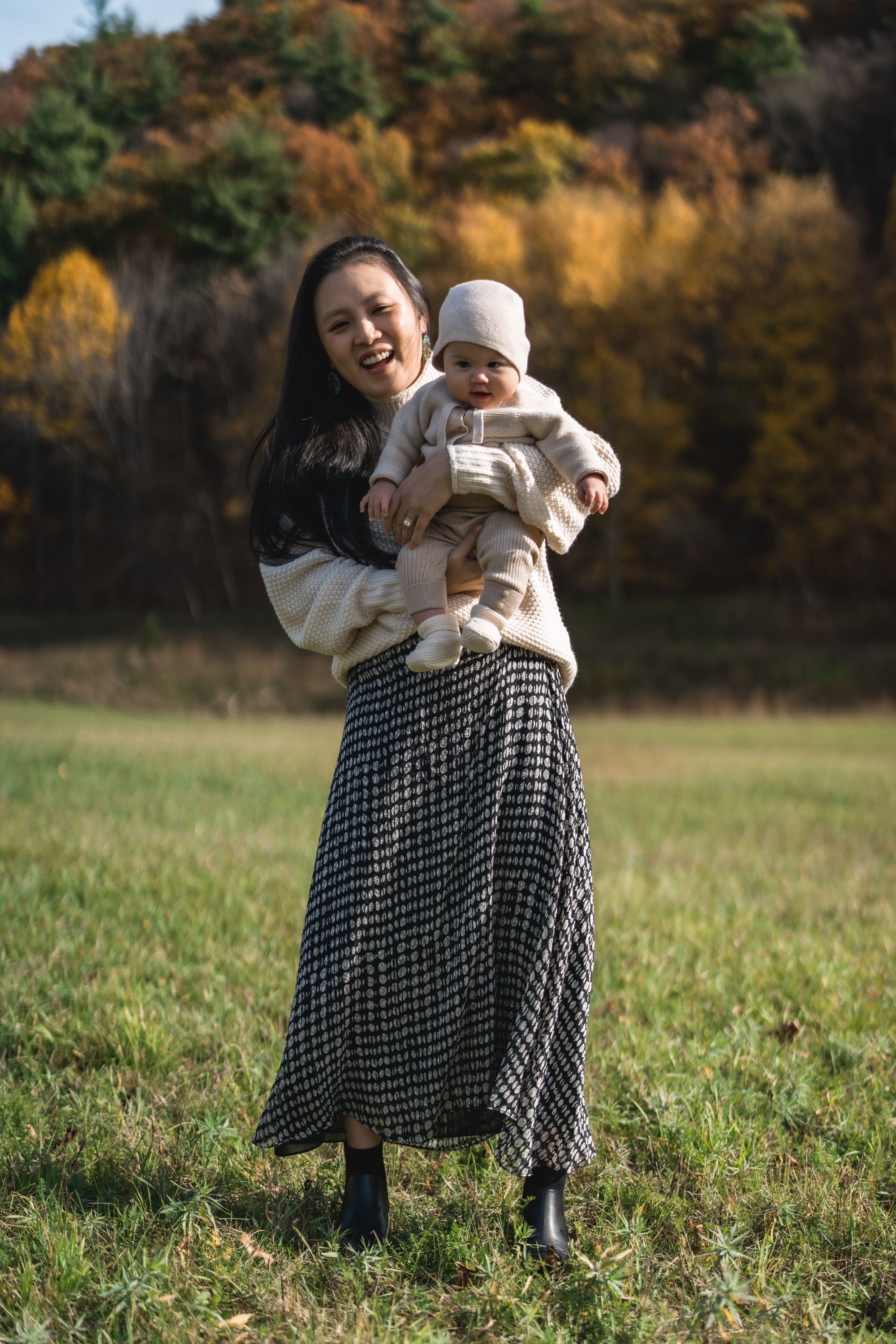 A woman holding an infant stands on a grassy field, smiling at the camera with autumn foliage and trees in soft focus behind them