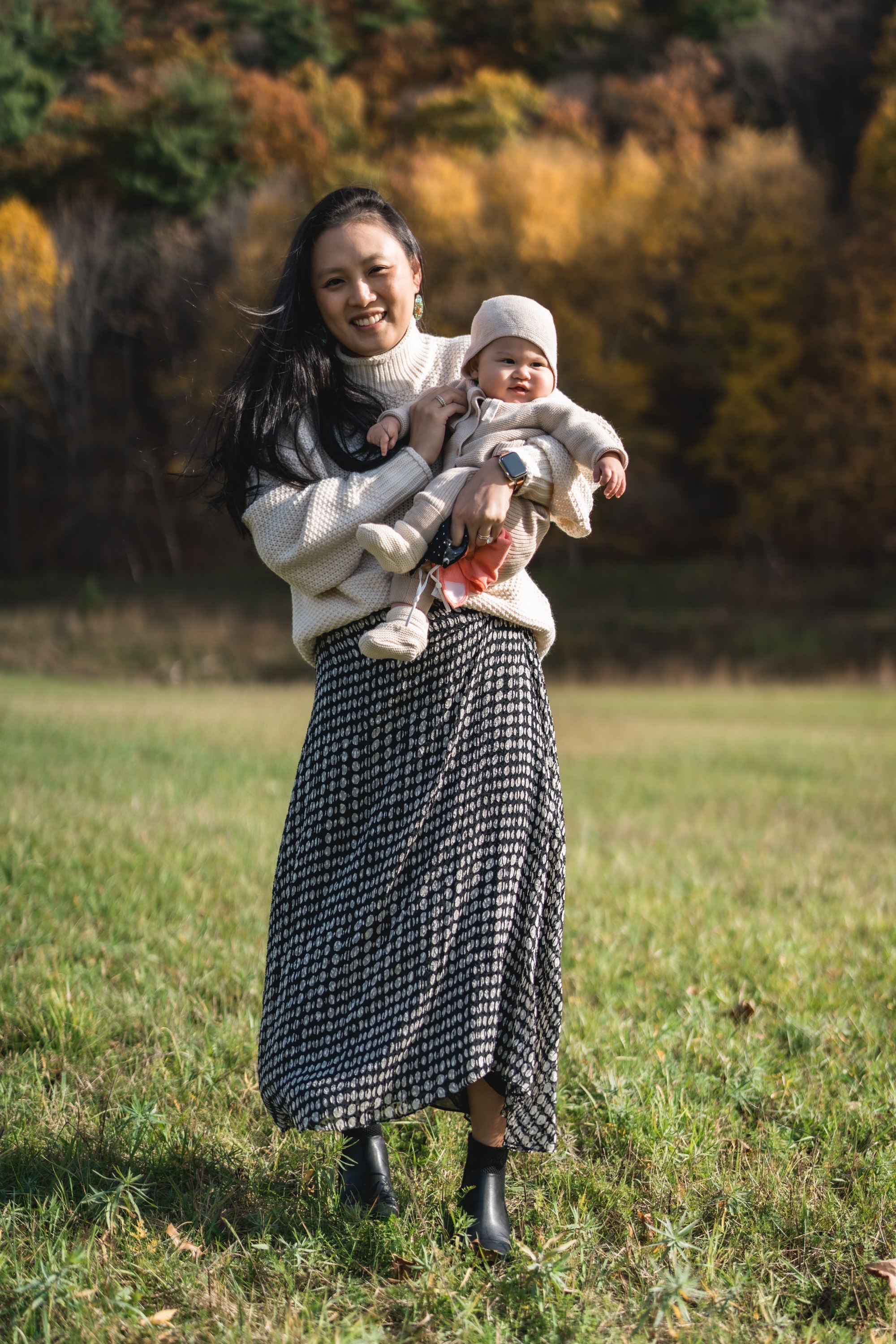 Woman in cream sweater and patterned skirt holds smiling infant in white knit outfit on grass, with golden autumn foliage blurred in background