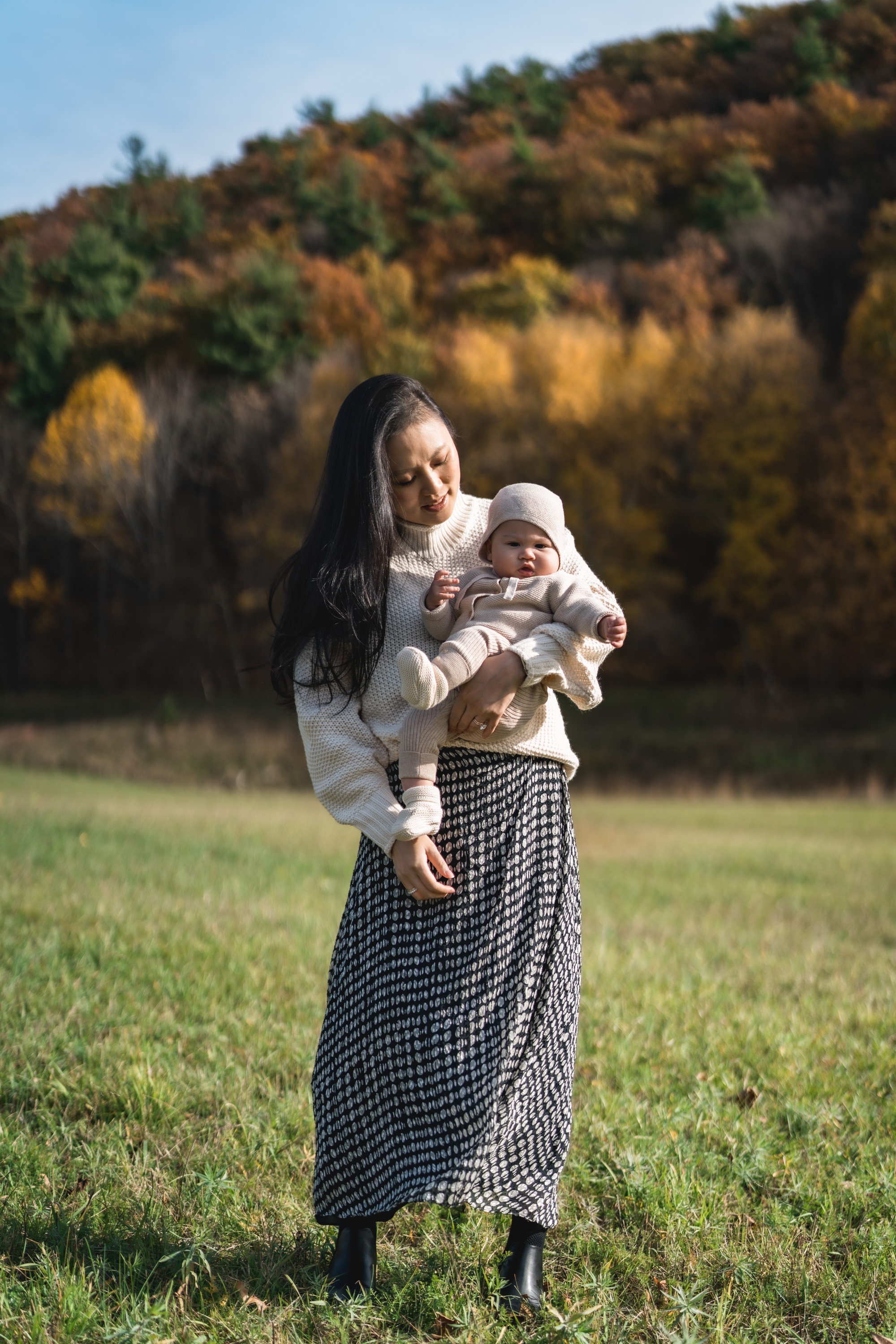 Woman holding a bundled infant stands in a grassy meadow beneath a forested hillside displaying autumn foliage in shades of gold, orange, and green