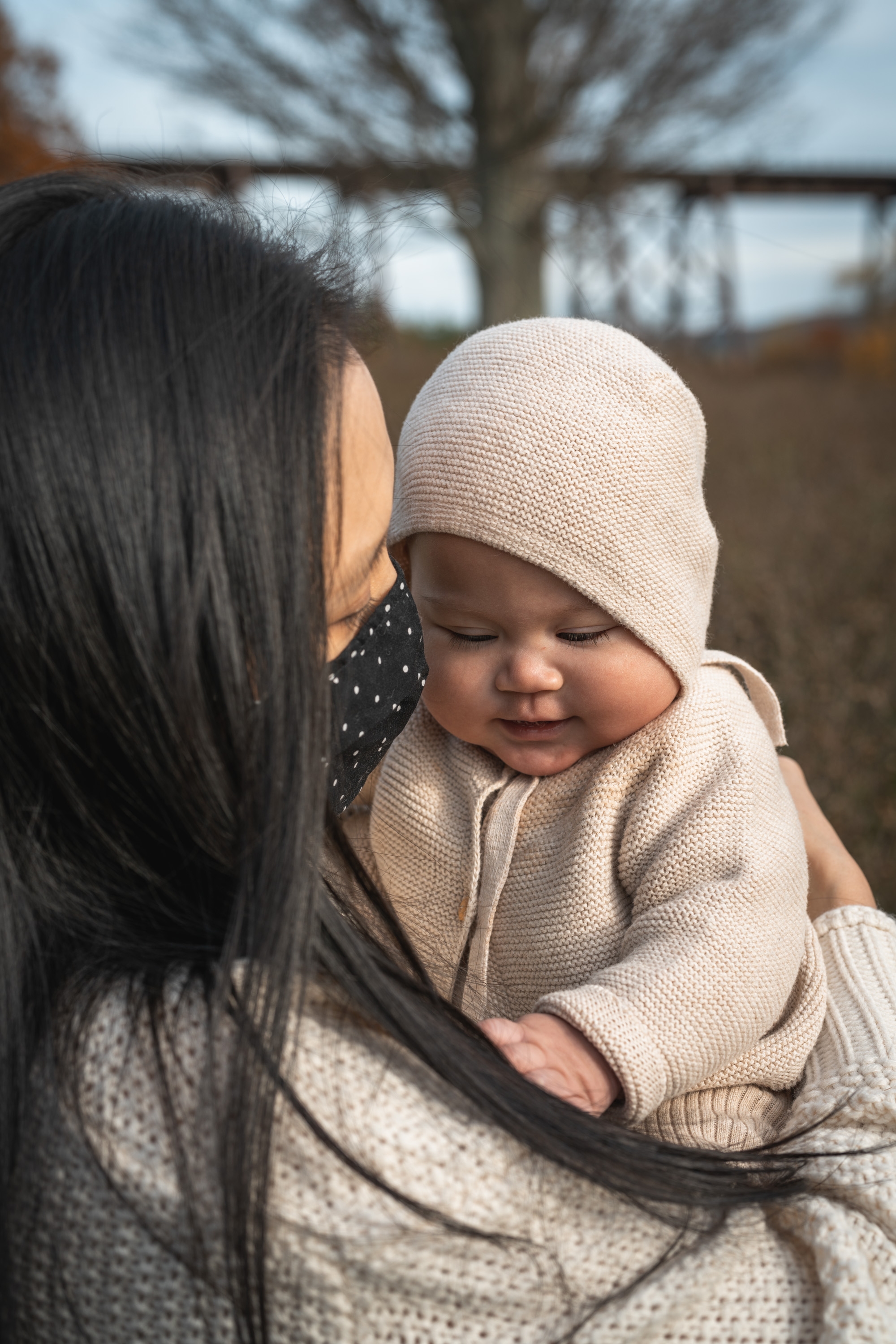 A woman with long black hair holds a smiling infant dressed in a cream knit beanie and sweater, with bare autumn trees and overcast sky visible behind them