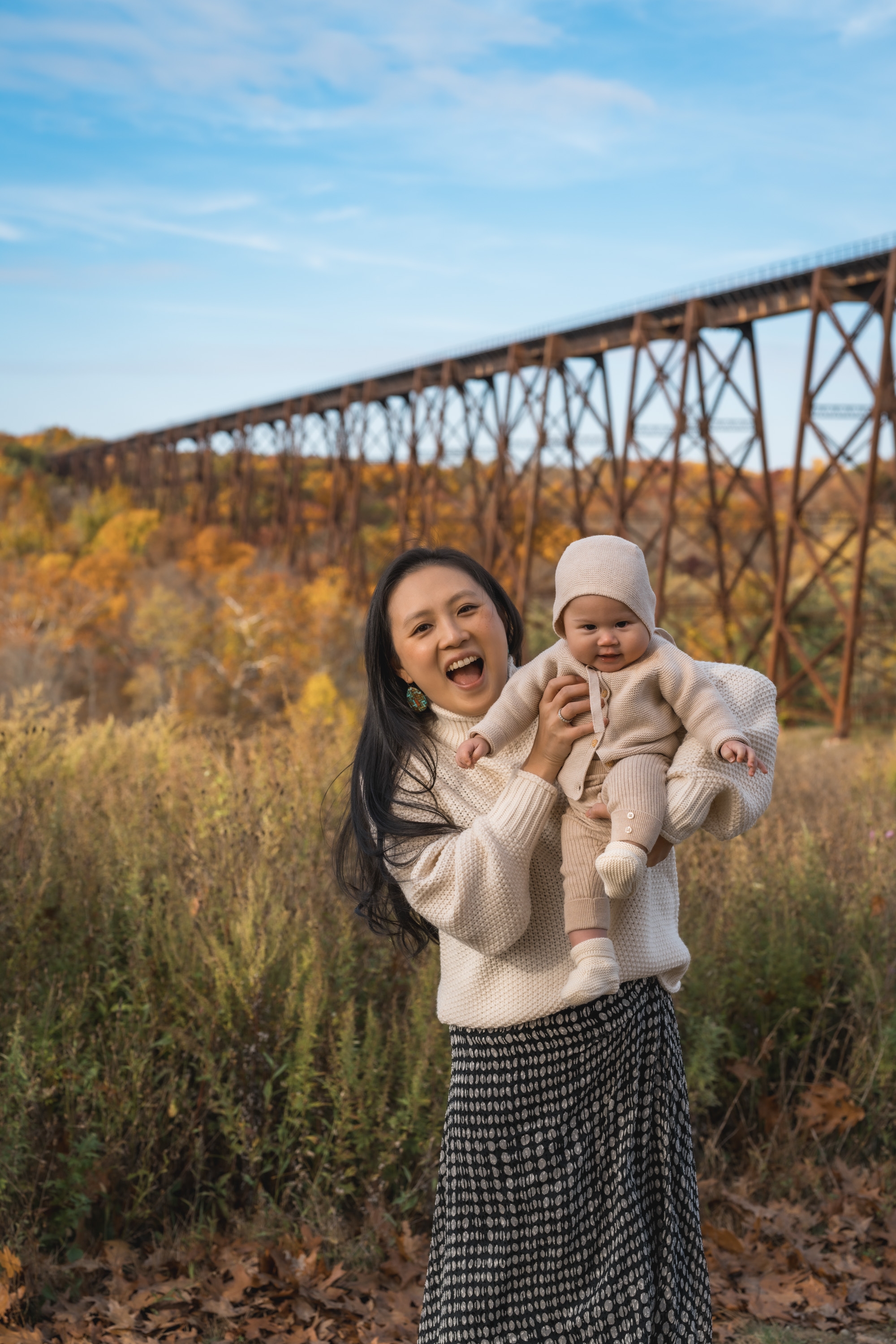A woman in a cream sweater holds a smiling infant in matching knit clothes, standing in golden autumn foliage with an iron trestle bridge spanning the valley behind them