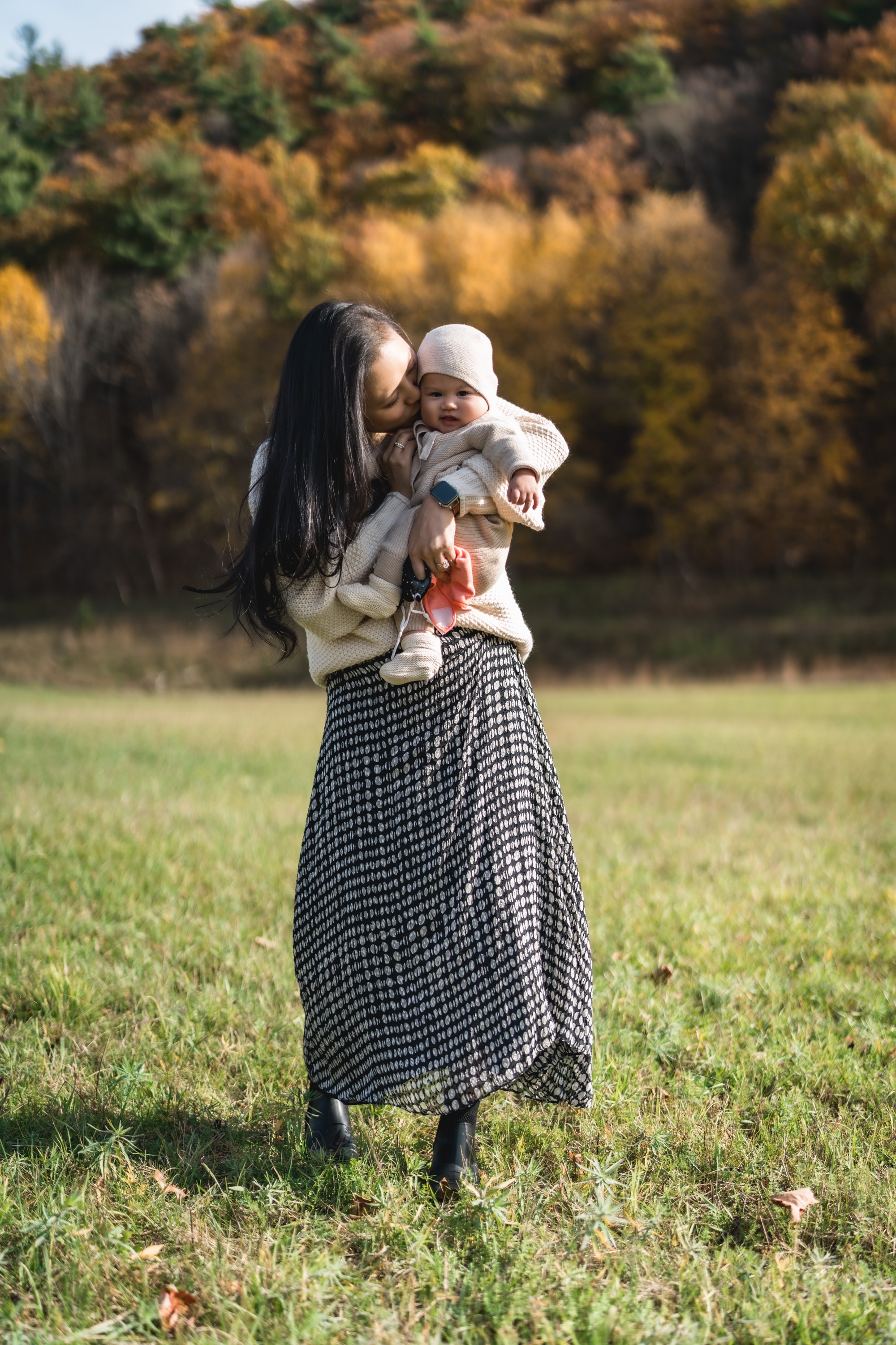 A woman holds her infant child in an open grassy field with autumn foliage in soft focus behind them