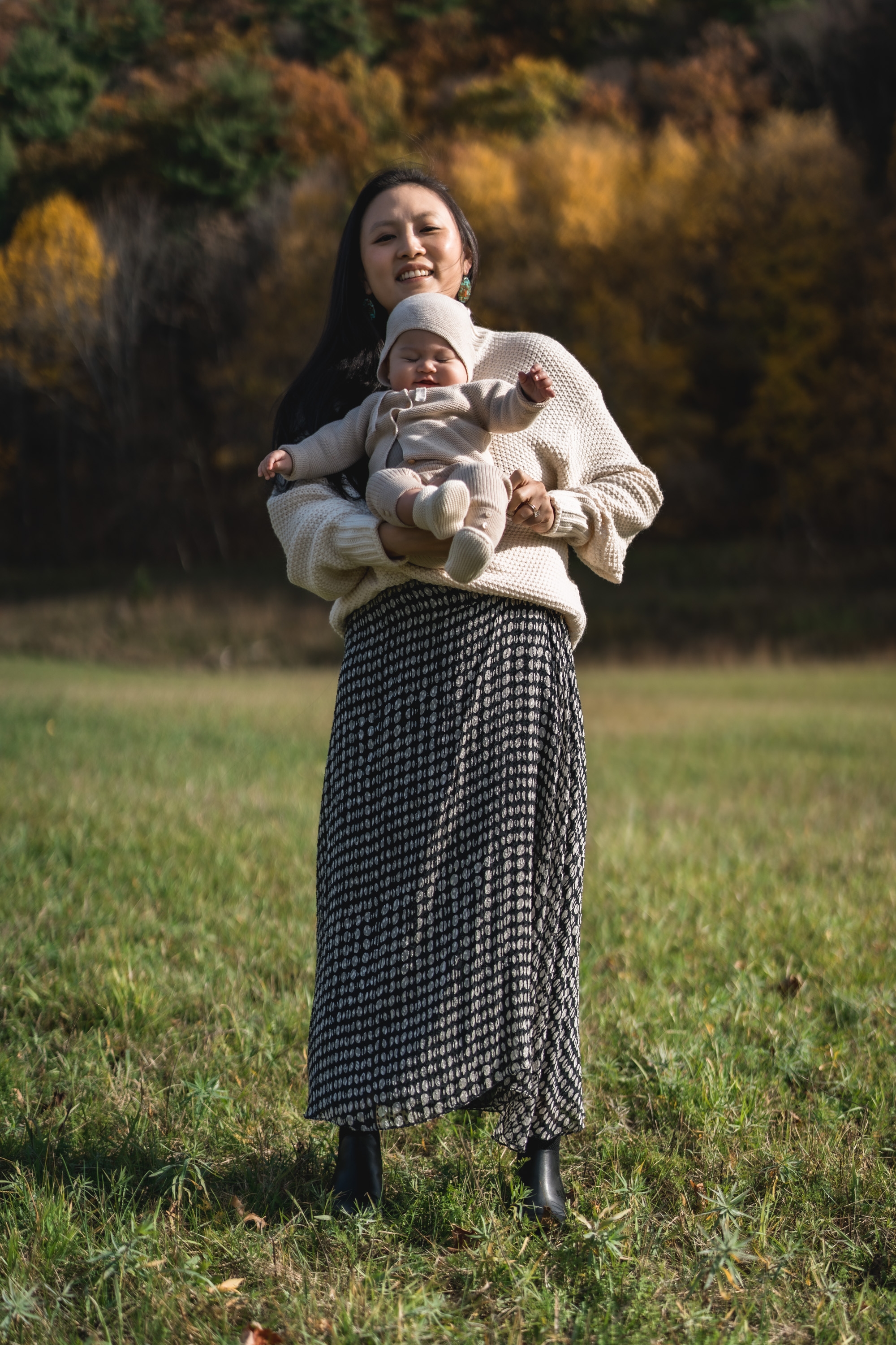 A woman with long dark hair holds her infant daughter in a cream-colored sweater while standing in a grassy field with autumn foliage glowing softly in the blurred background