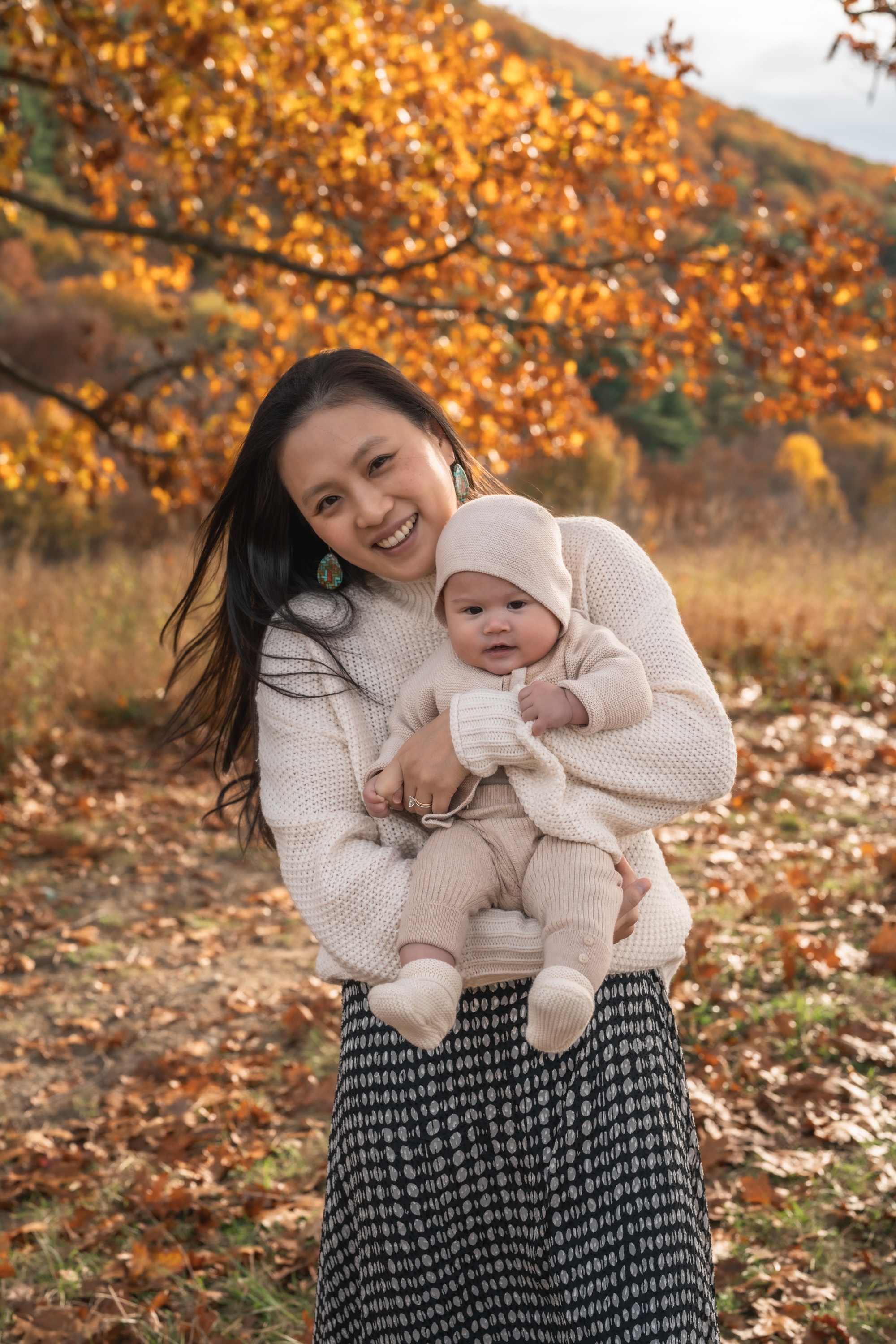 Woman holding a bundled infant in cream knit clothing stands smiling before a hillside ablaze with golden autumn foliage and scattered fallen leaves