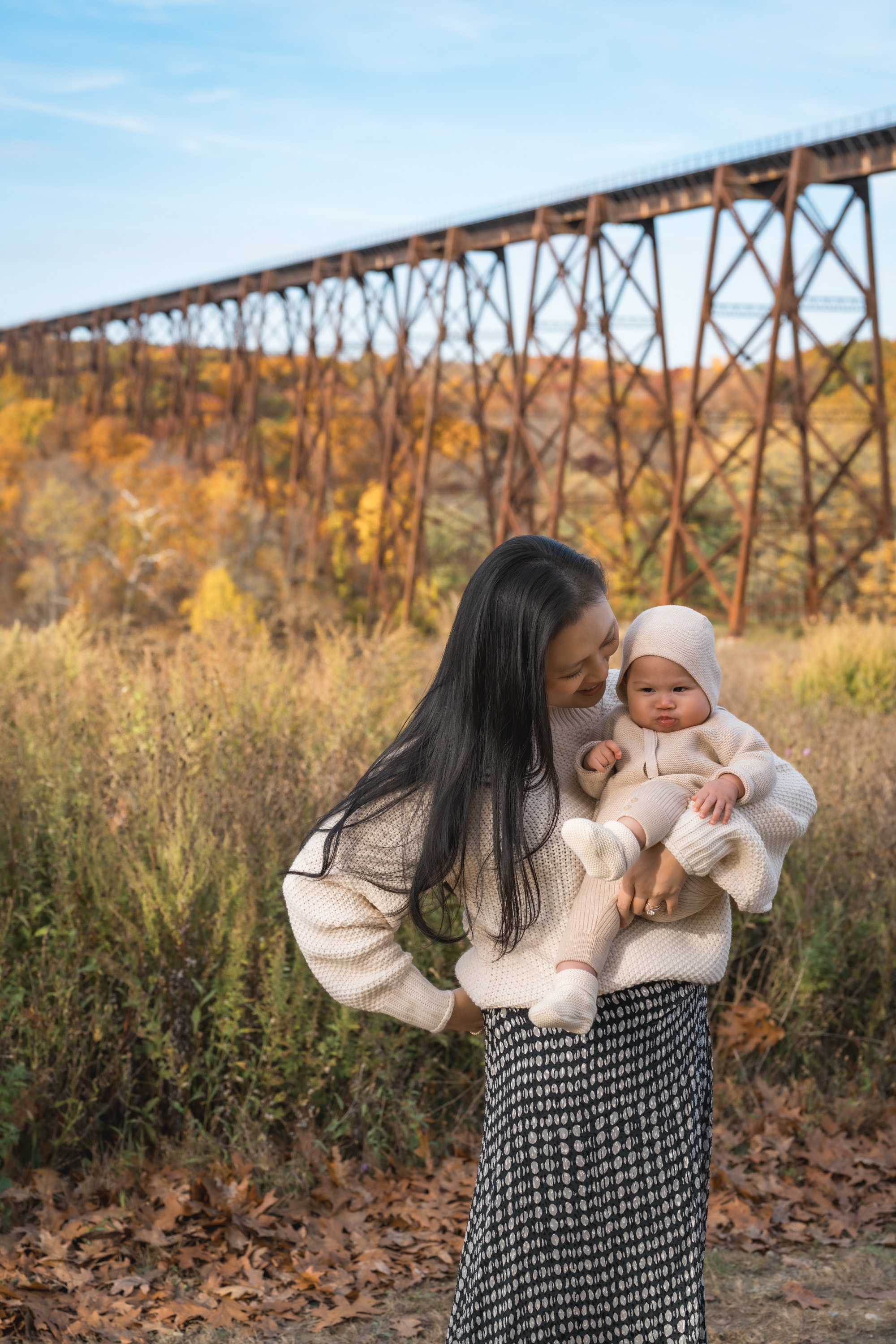 A woman holds an infant in cream-colored clothing against a backdrop of golden autumn foliage and an industrial steel trestle bridge spanning a forested valley
