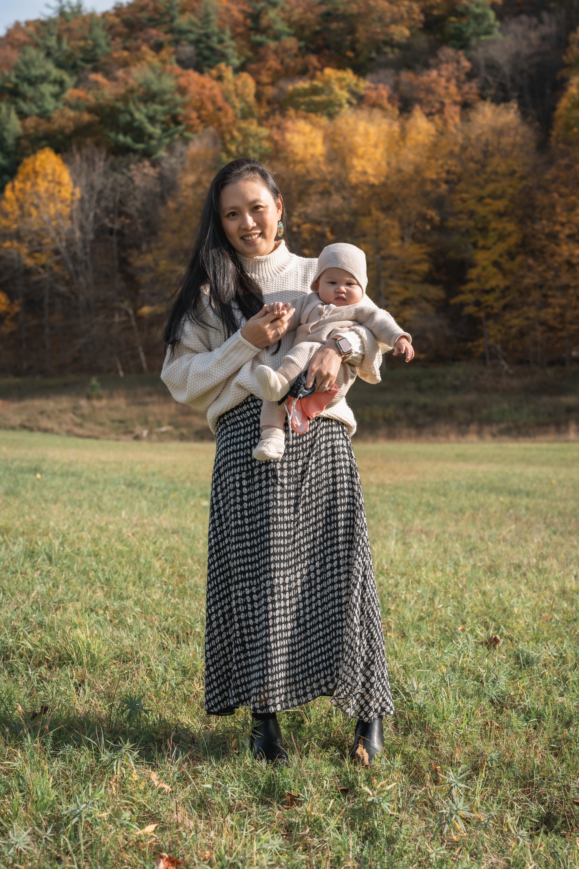A smiling mother holds her infant child in a cream-colored knit sweater while standing in an open grass field with blurred autumn foliage behind them