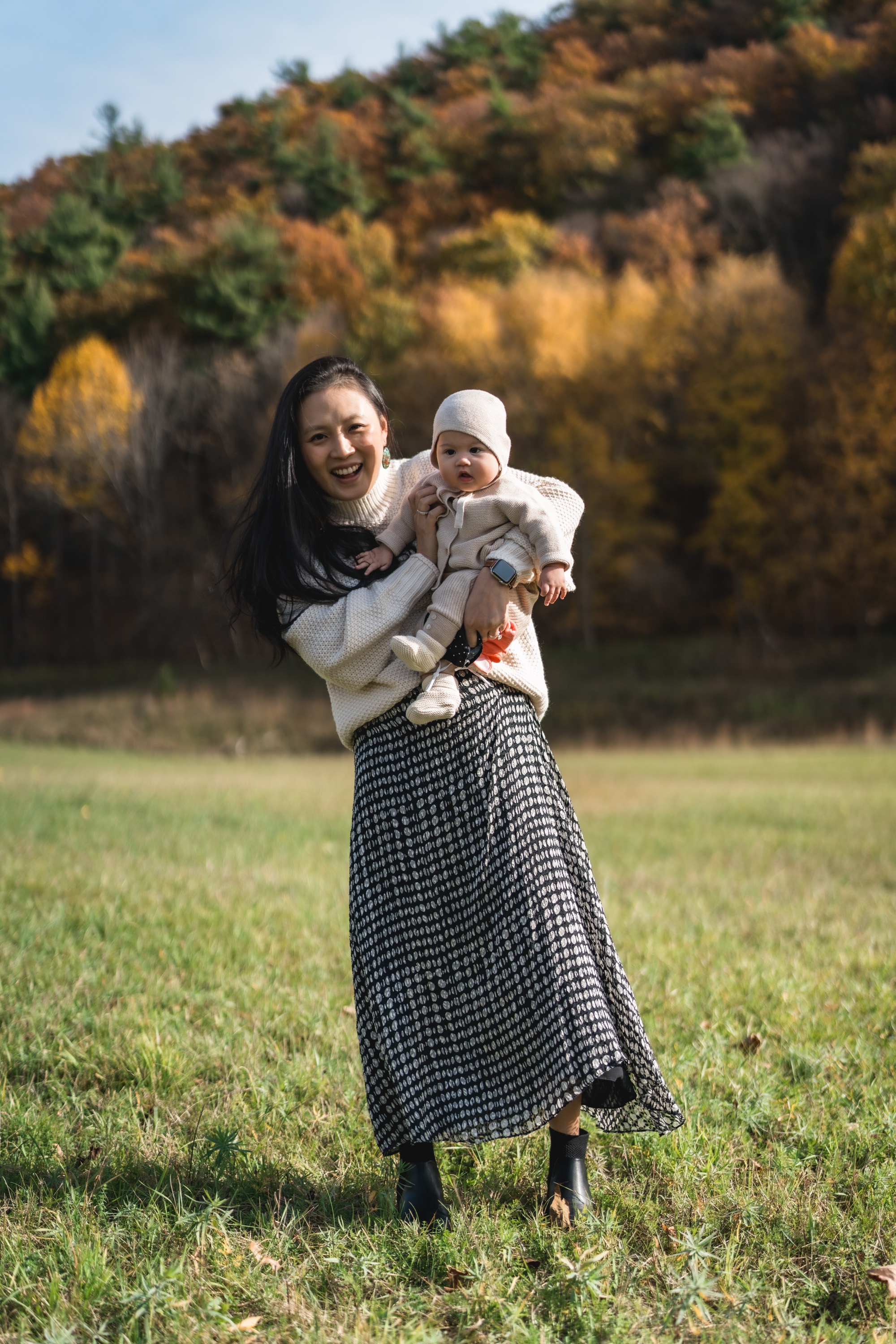 A woman holds her infant child in a grassy meadow beneath autumn foliage, with golden and green trees creating a warm backdrop under clear daylight