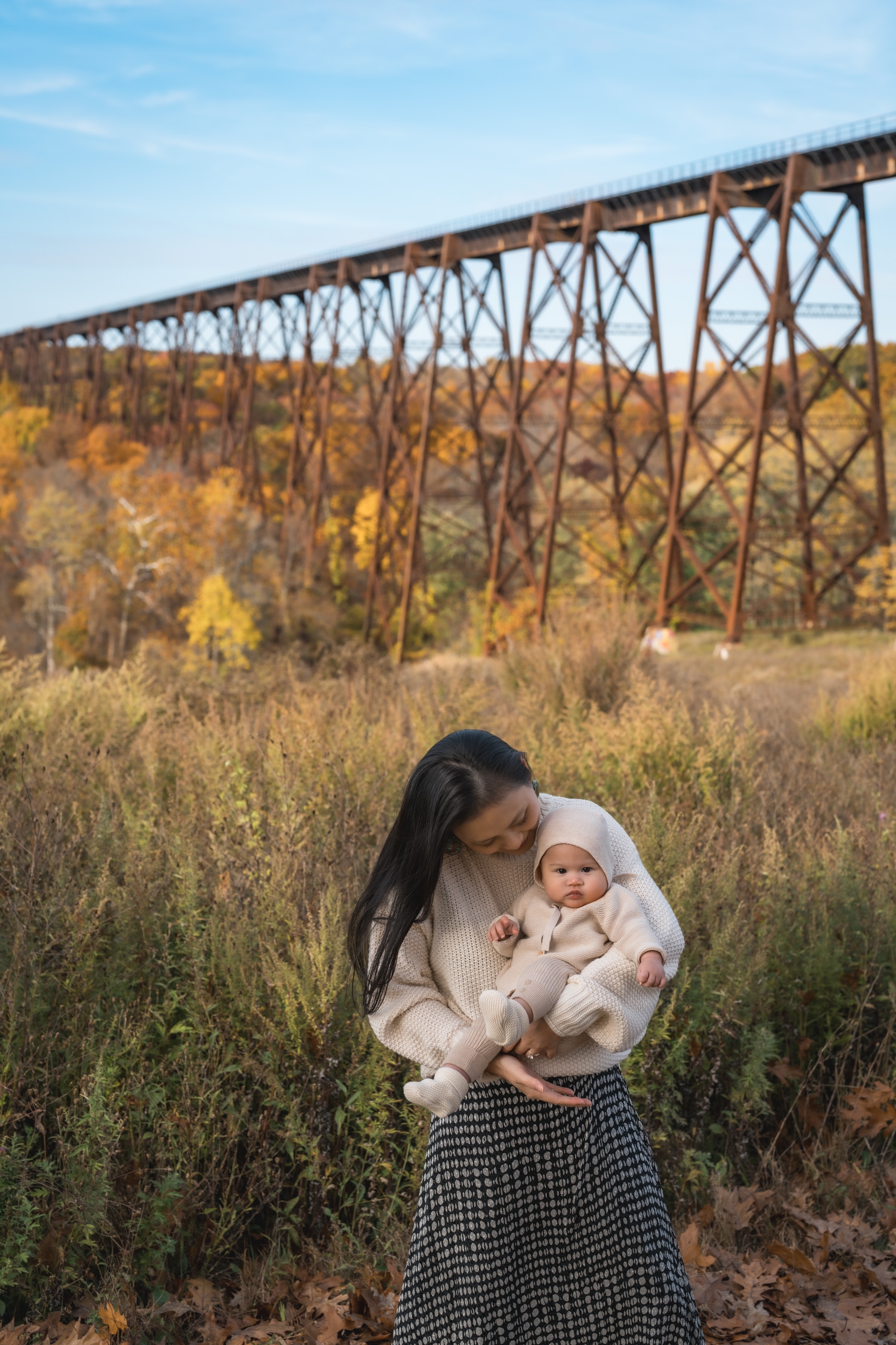 Woman holding an infant in a cream-colored sweater stands in golden autumn grassland beneath a towering russet iron railroad trestle