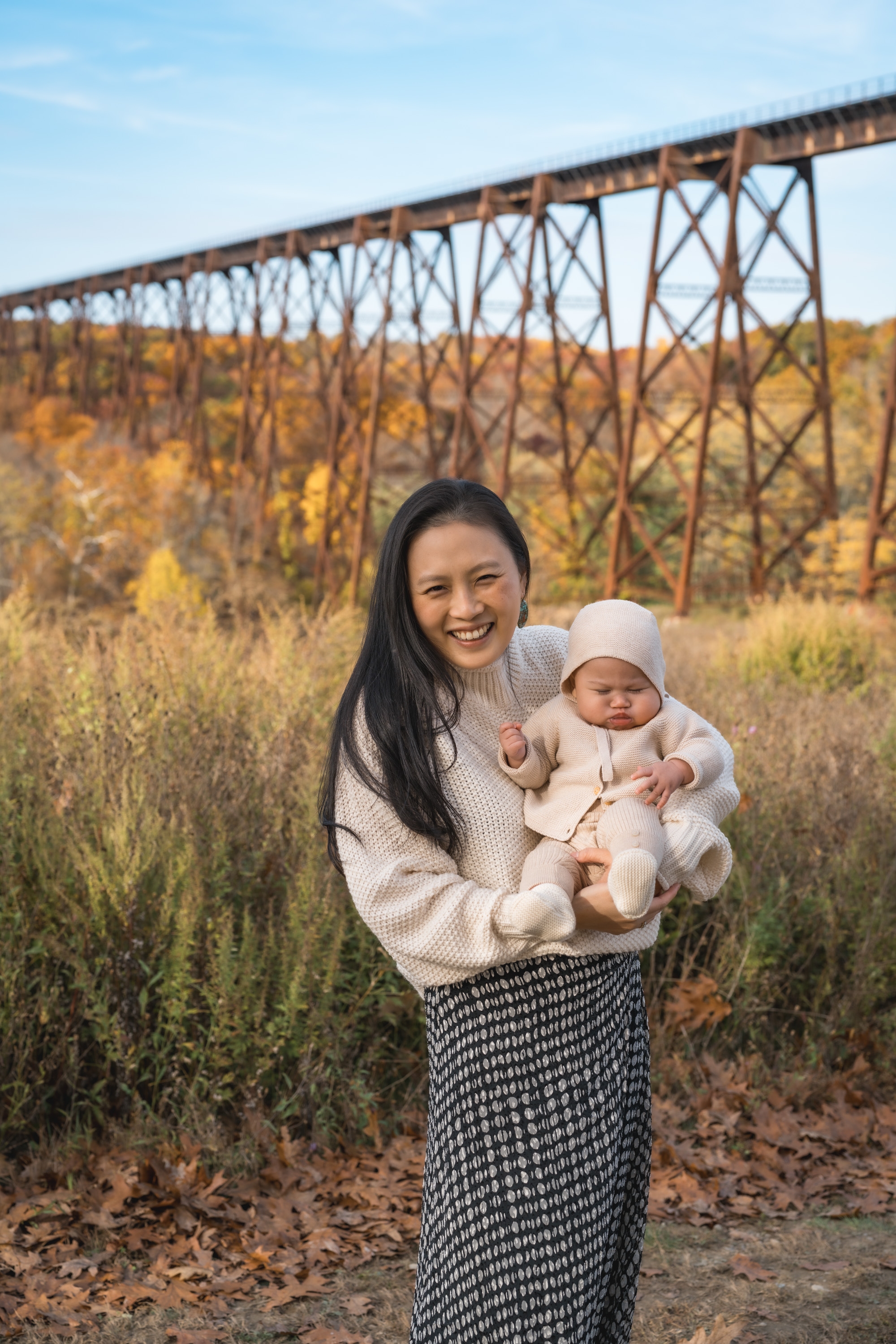 A woman in a cream sweater holds her infant in white clothing while standing in golden autumn grassland beneath a tall metal railroad trestle bridge