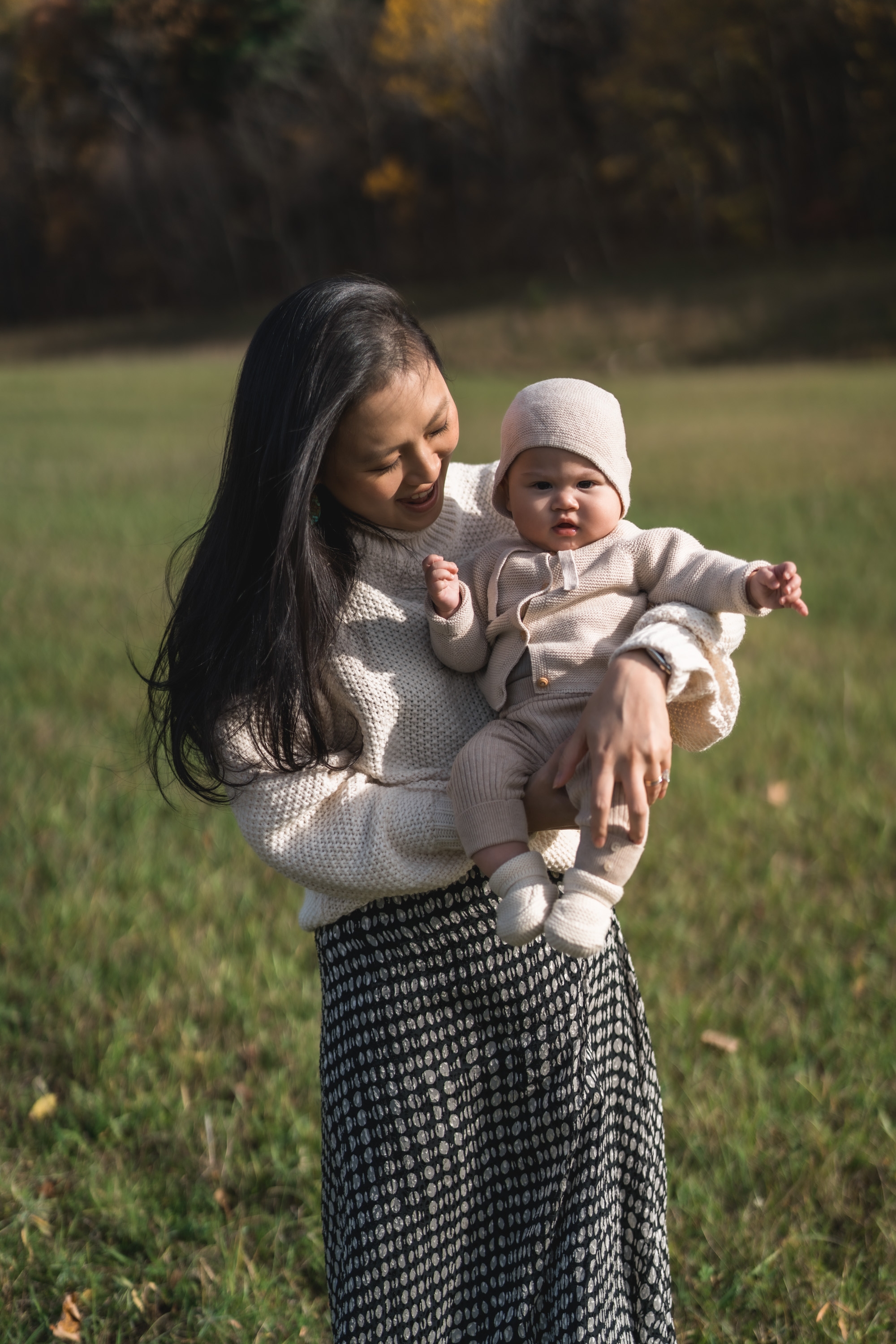 A woman with long dark hair holds a smiling infant wearing a cream-colored hat and sweater in an open grassy field with autumn foliage blurred in the background