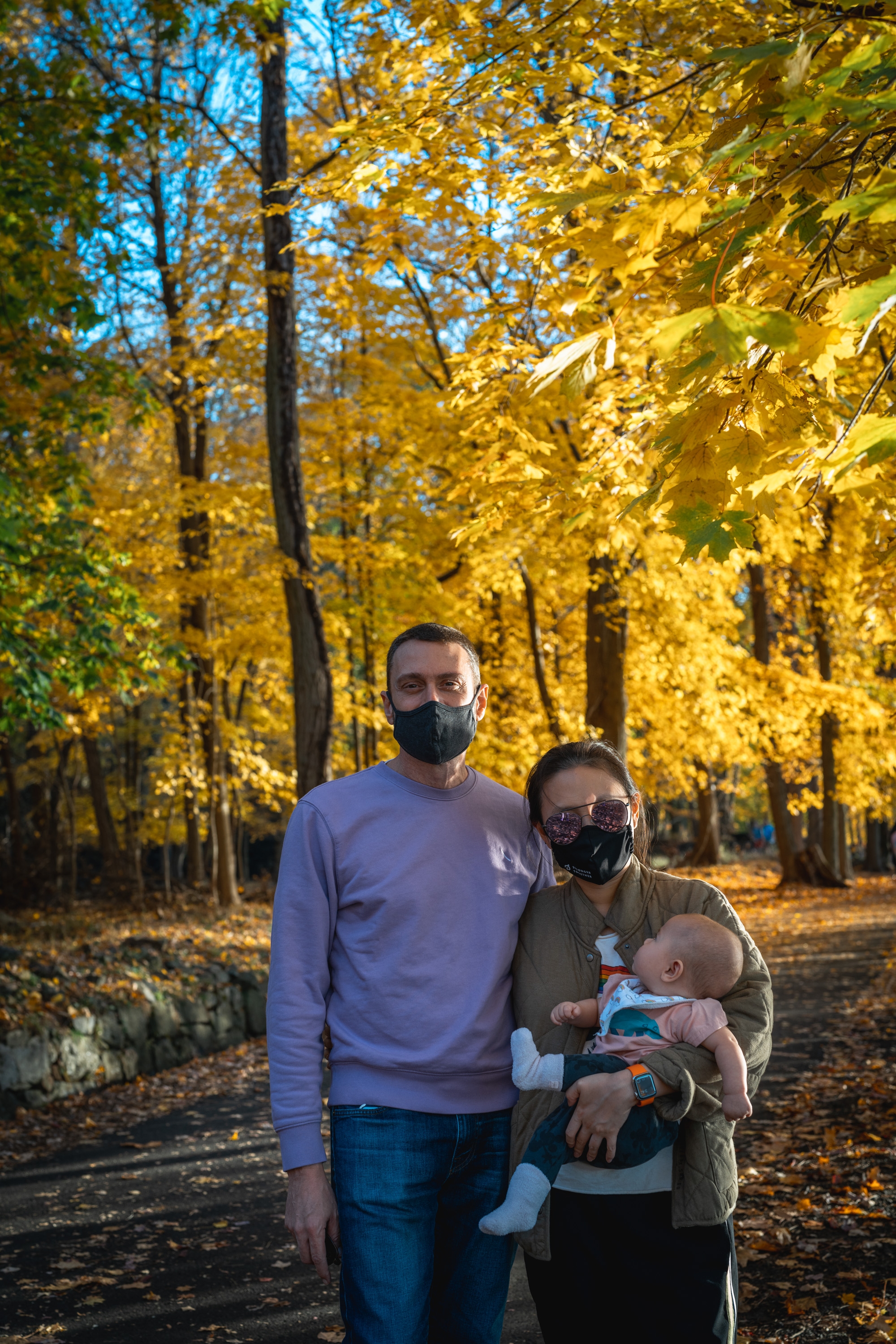 A family of three wearing masks stands on a leaf-covered path surrounded by golden autumn trees under clear blue sky