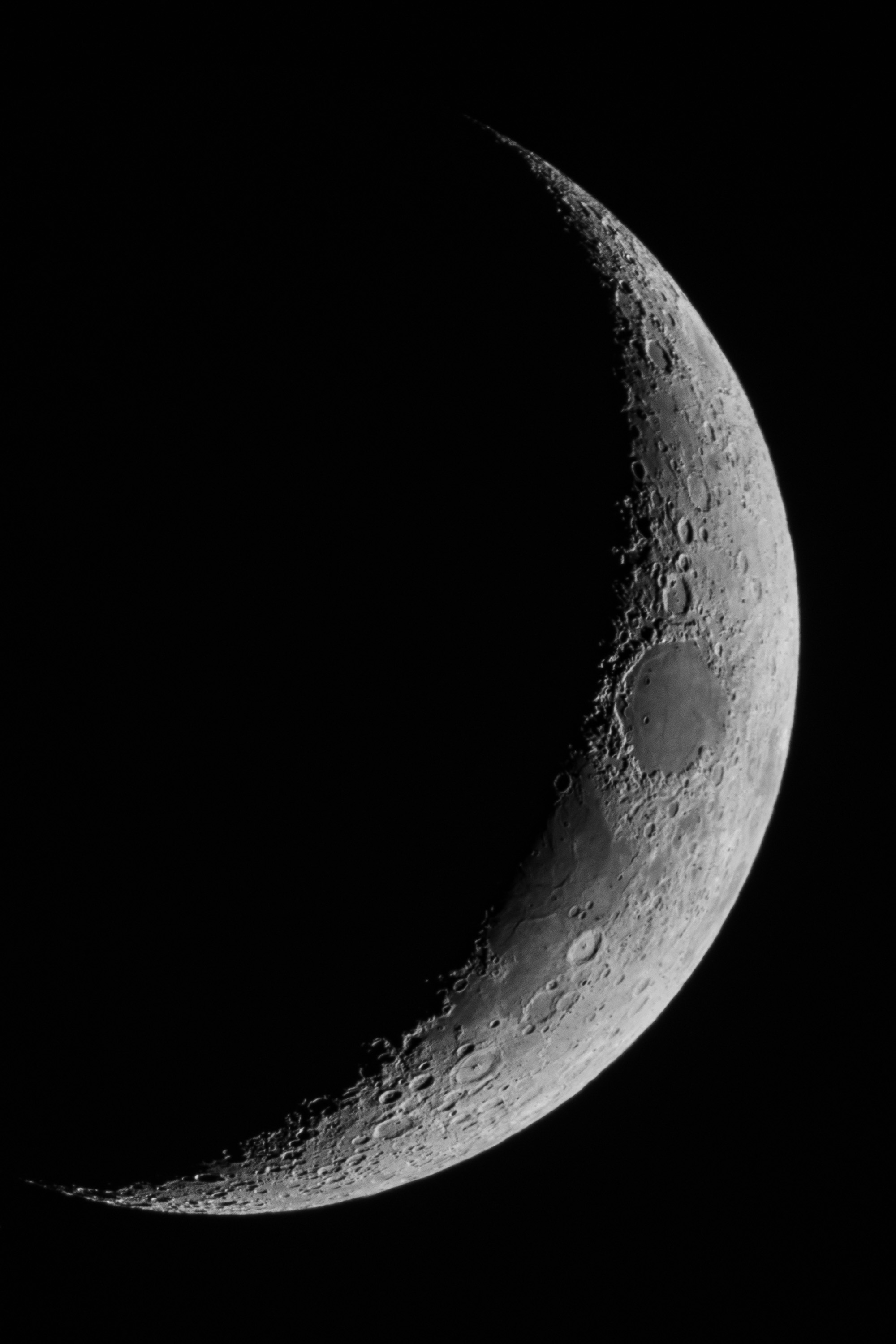 A crescent moon captured against a black night sky reveals detailed lunar craters and surface texture along its illuminated edge