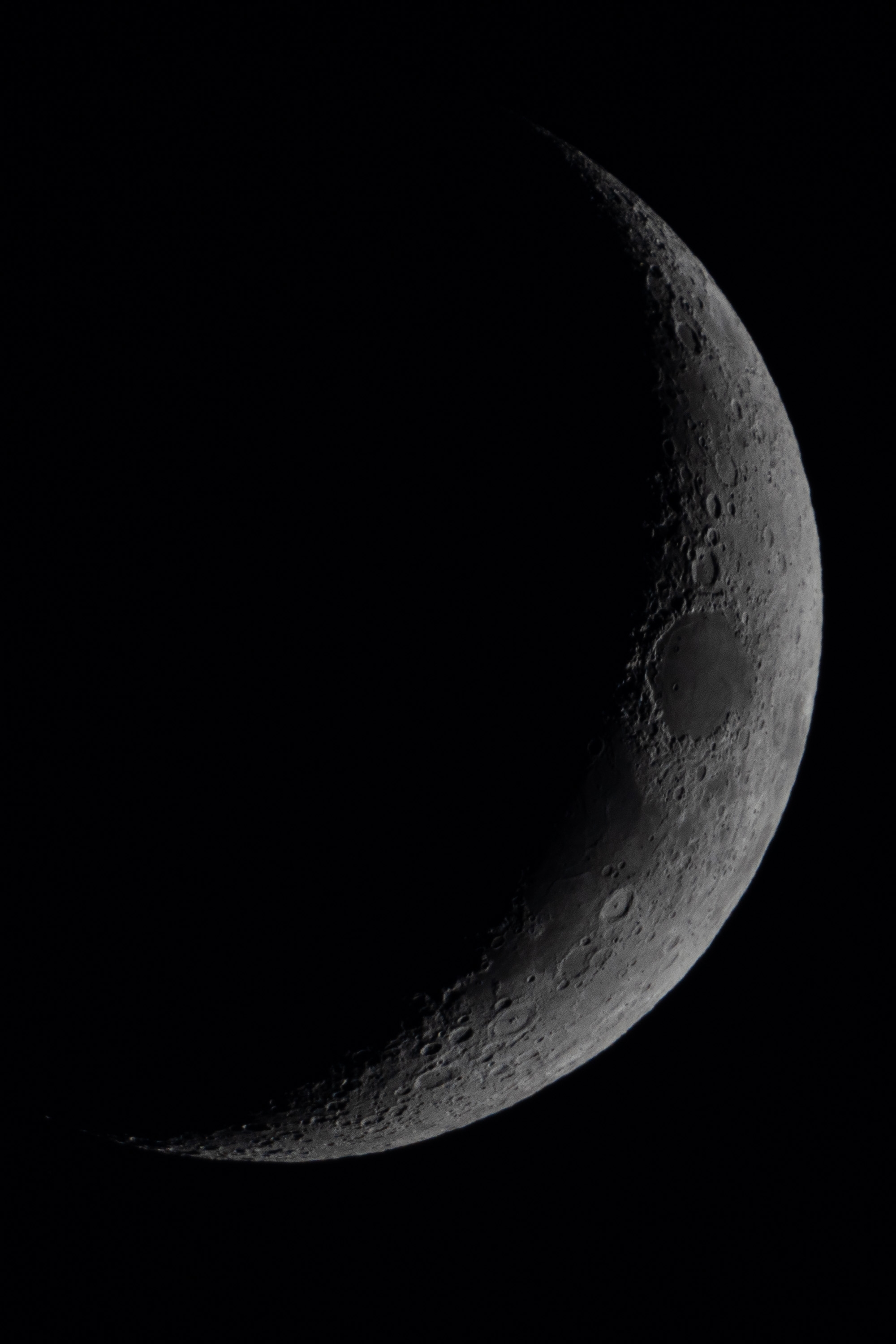 A crescent moon with detailed surface features and crater formations illuminated against a black night sky, captured with a zoom lens revealing textured lunar terrain along the terminator line