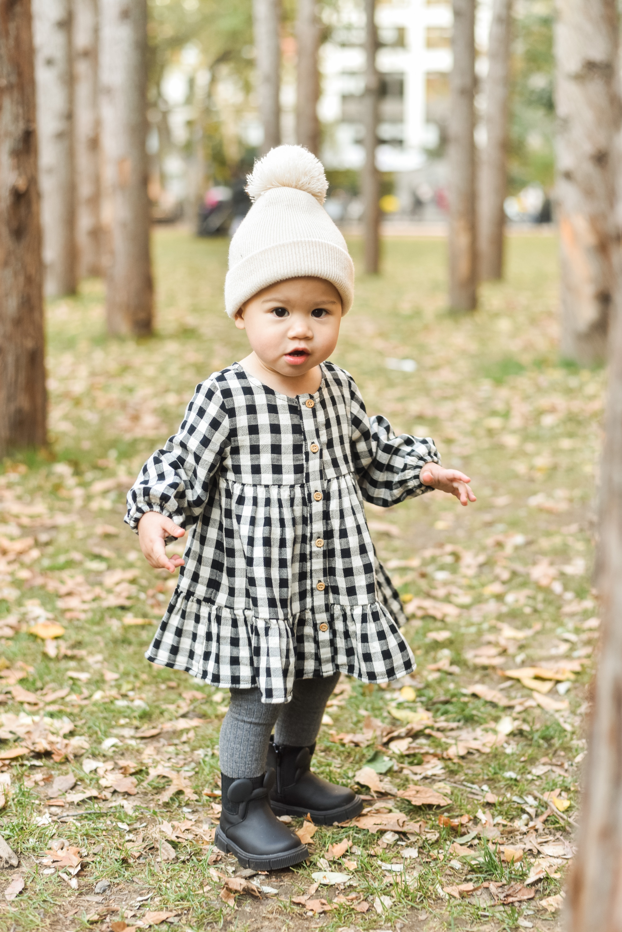 Toddler in a white pom-pom beanie and black-and-white gingham dress stands on a tree-lined path scattered with fallen leaves, with a cemetery cross visible in the soft-focus background
