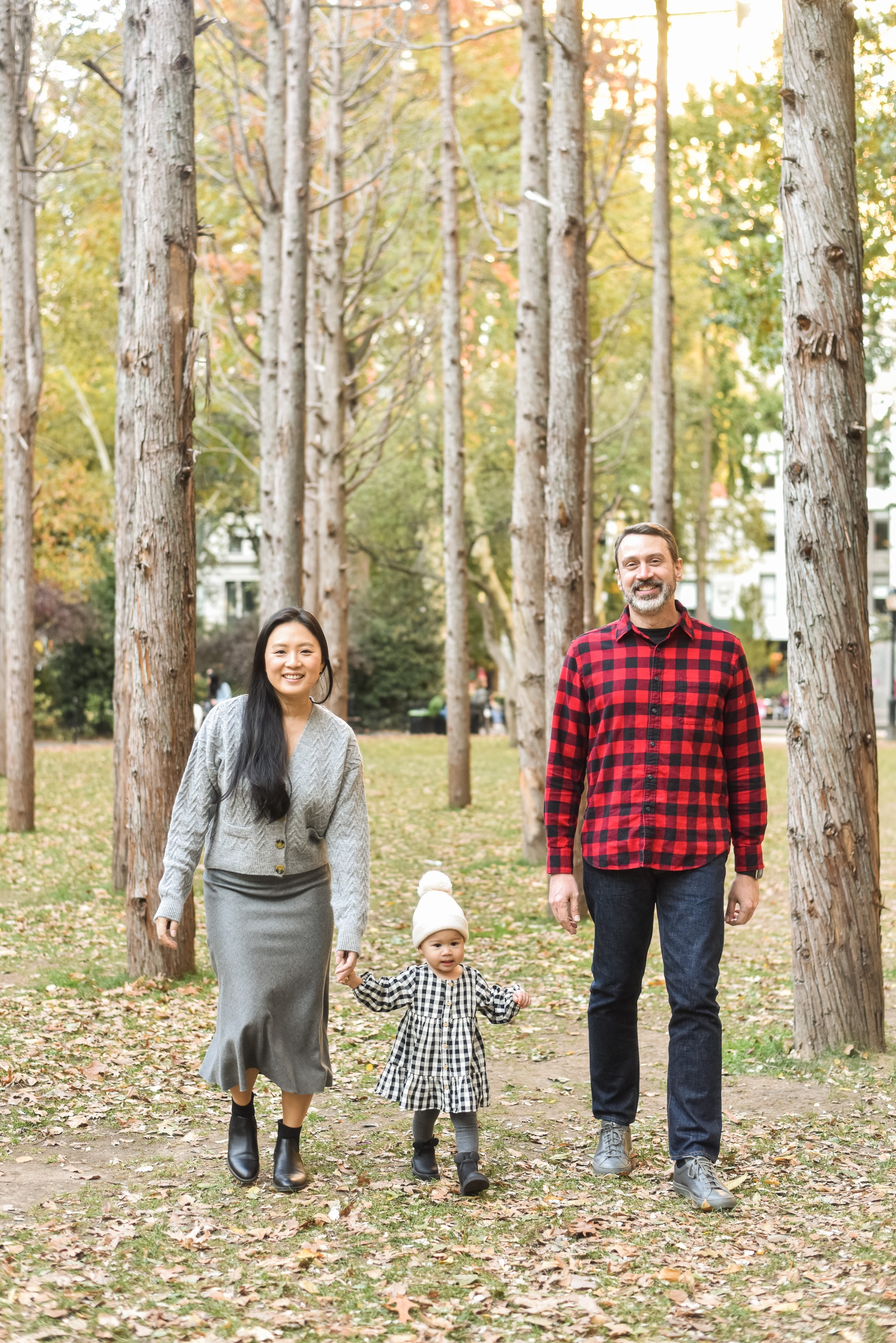 A family of three walks hand-in-hand down a tree-lined path covered with fallen leaves during autumn, with tall birch trees framing the composition
