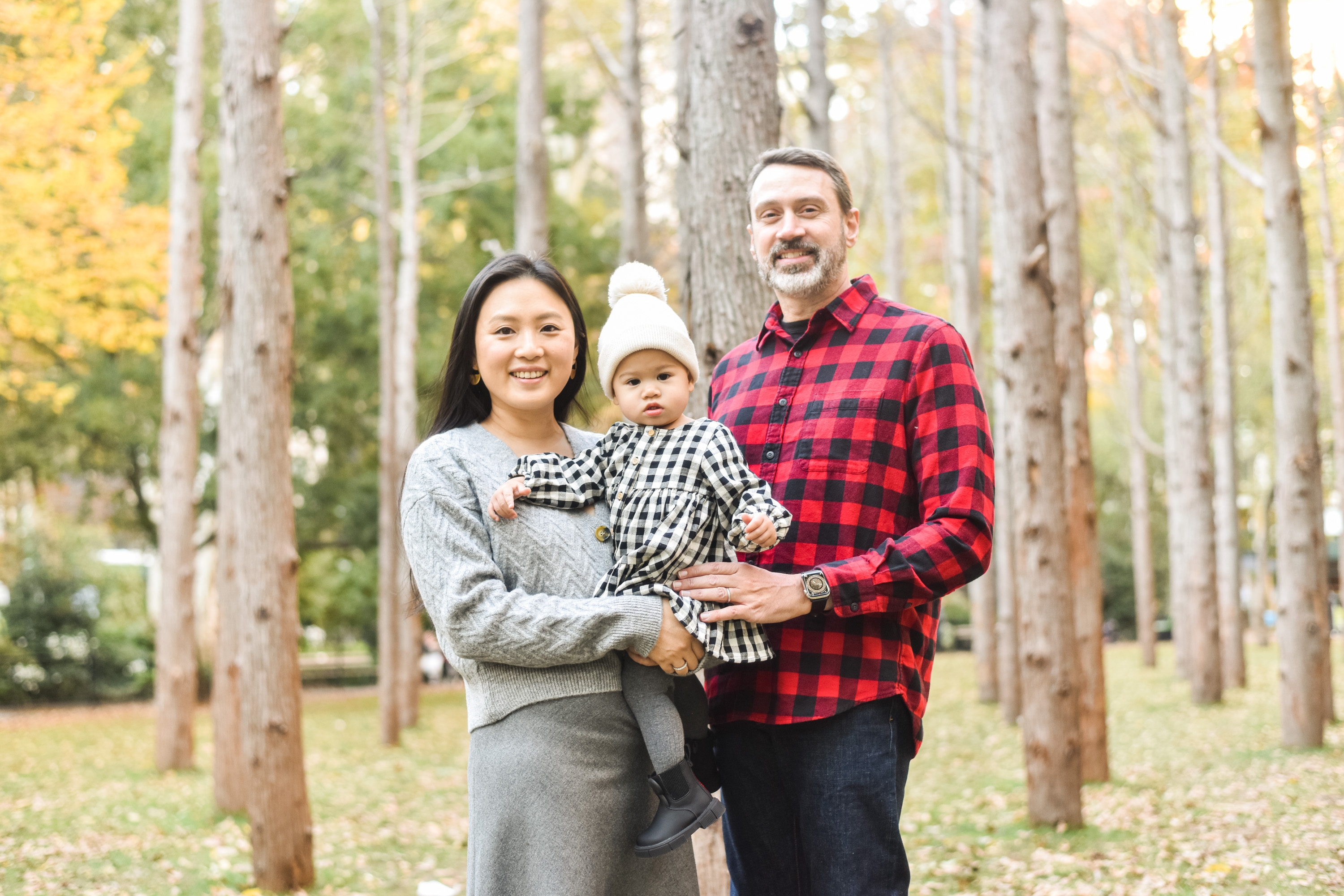 A family of three stands together among tall trees in autumn, with the mother in gray, father in red plaid, and toddler in white beanie positioned between them