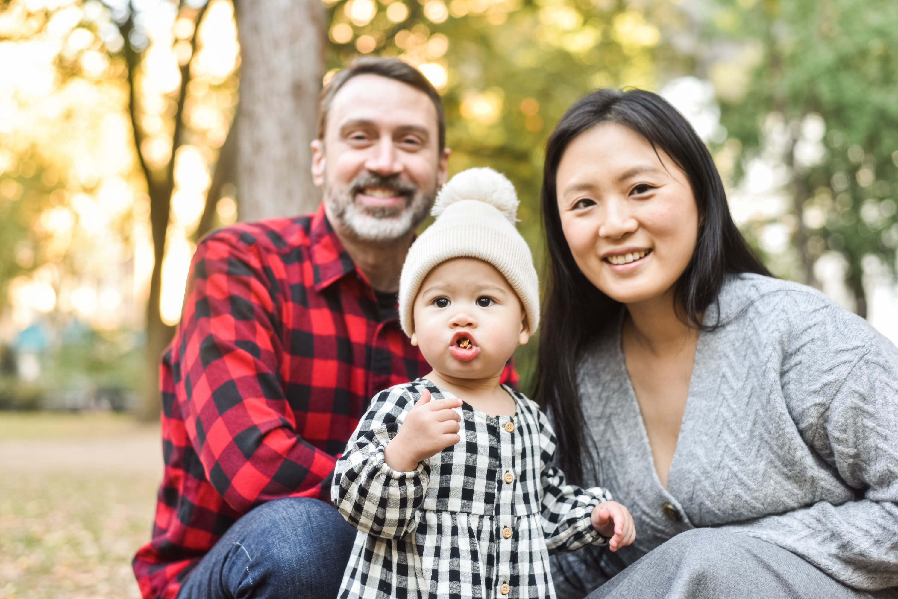 A smiling family of three—a bearded man in red plaid, a woman in gray cardigan, and a toddler in a white hat and checkered dress—pose together outdoors among autumn trees with soft golden-hour light filtering through the foliage