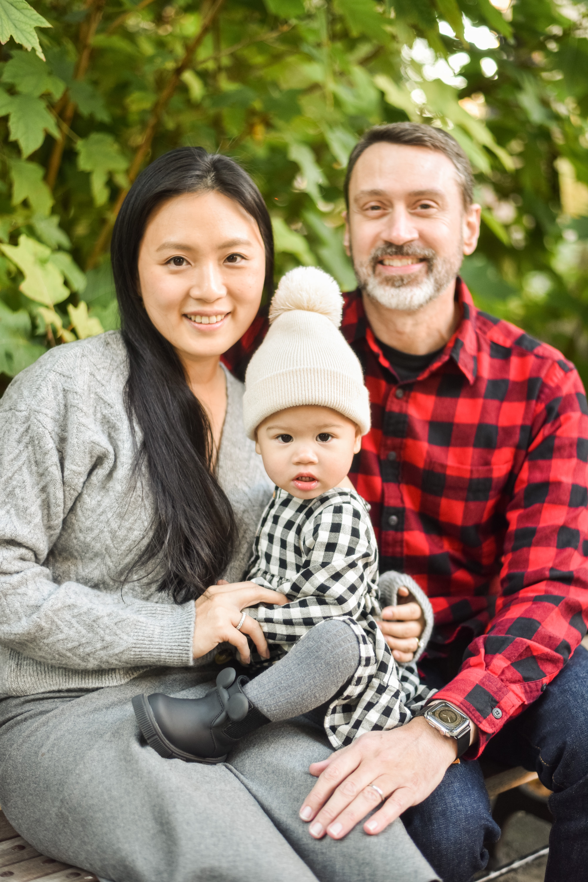 A young family of three smiles at the camera surrounded by lush green foliage, with the mother in gray, father in red plaid, and toddler wearing a cream beanie and checkered shirt