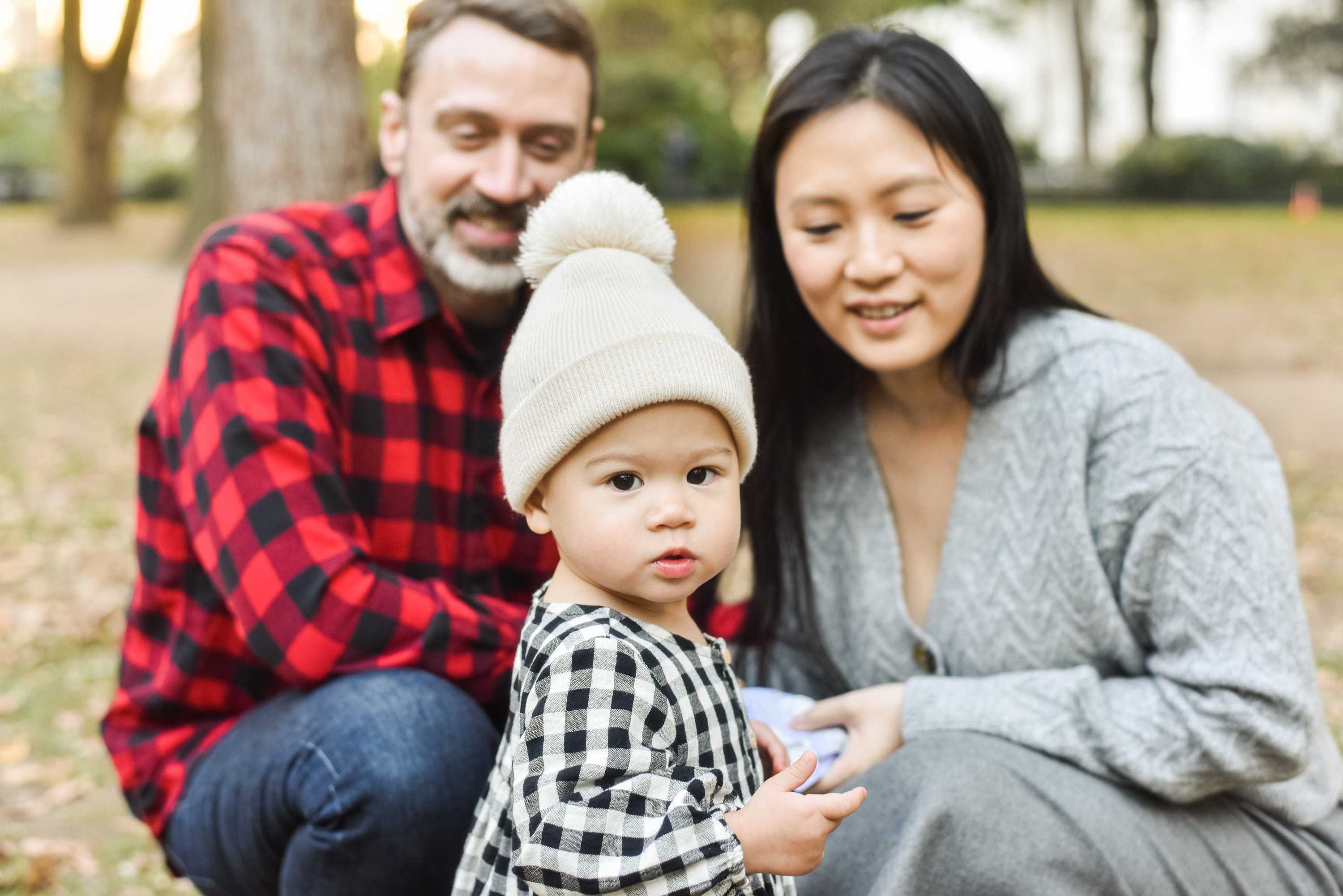 A young family of three poses outdoors in a park with tall trees, the toddler wearing a white knit beanie while parents in plaid and gray clothing smile nearby