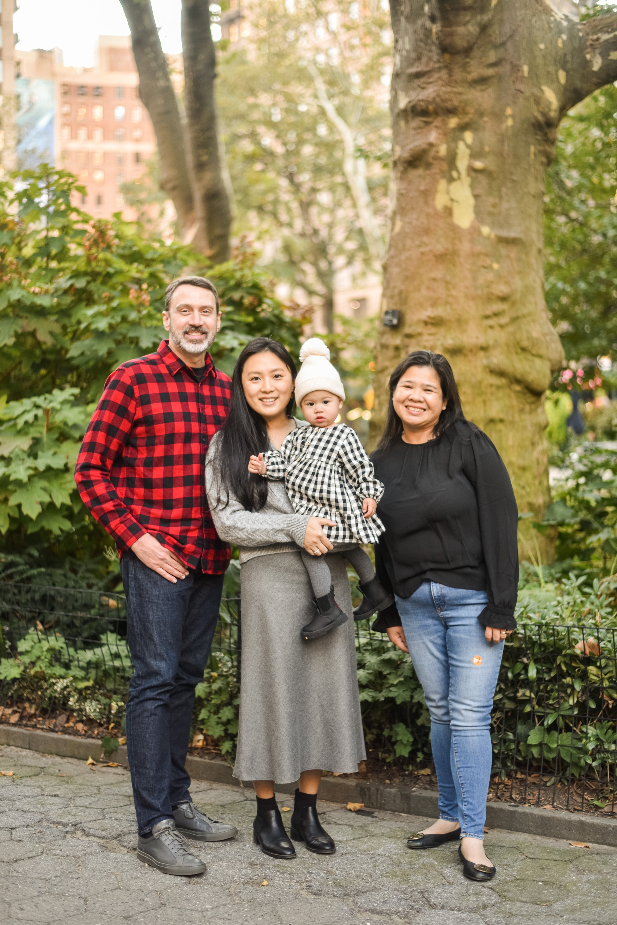 A family of four stands together smiling in a tree-lined urban garden, with brick buildings visible in the soft background and dappled sunlight filtering through the canopy above