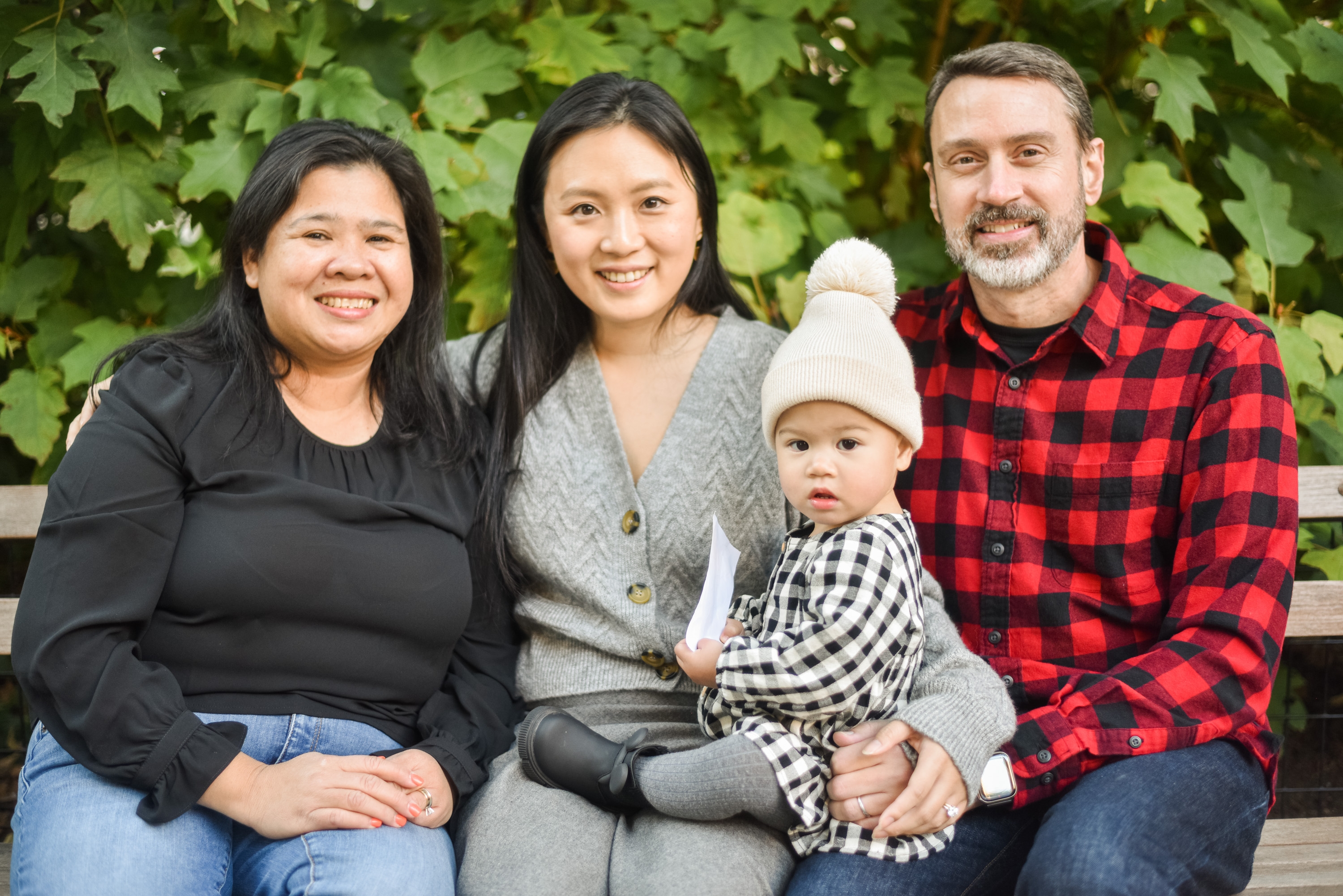 Four people sit on a wooden bench against lush green foliage, with a toddler in a white beanie centered between two women and a man in a red plaid shirt