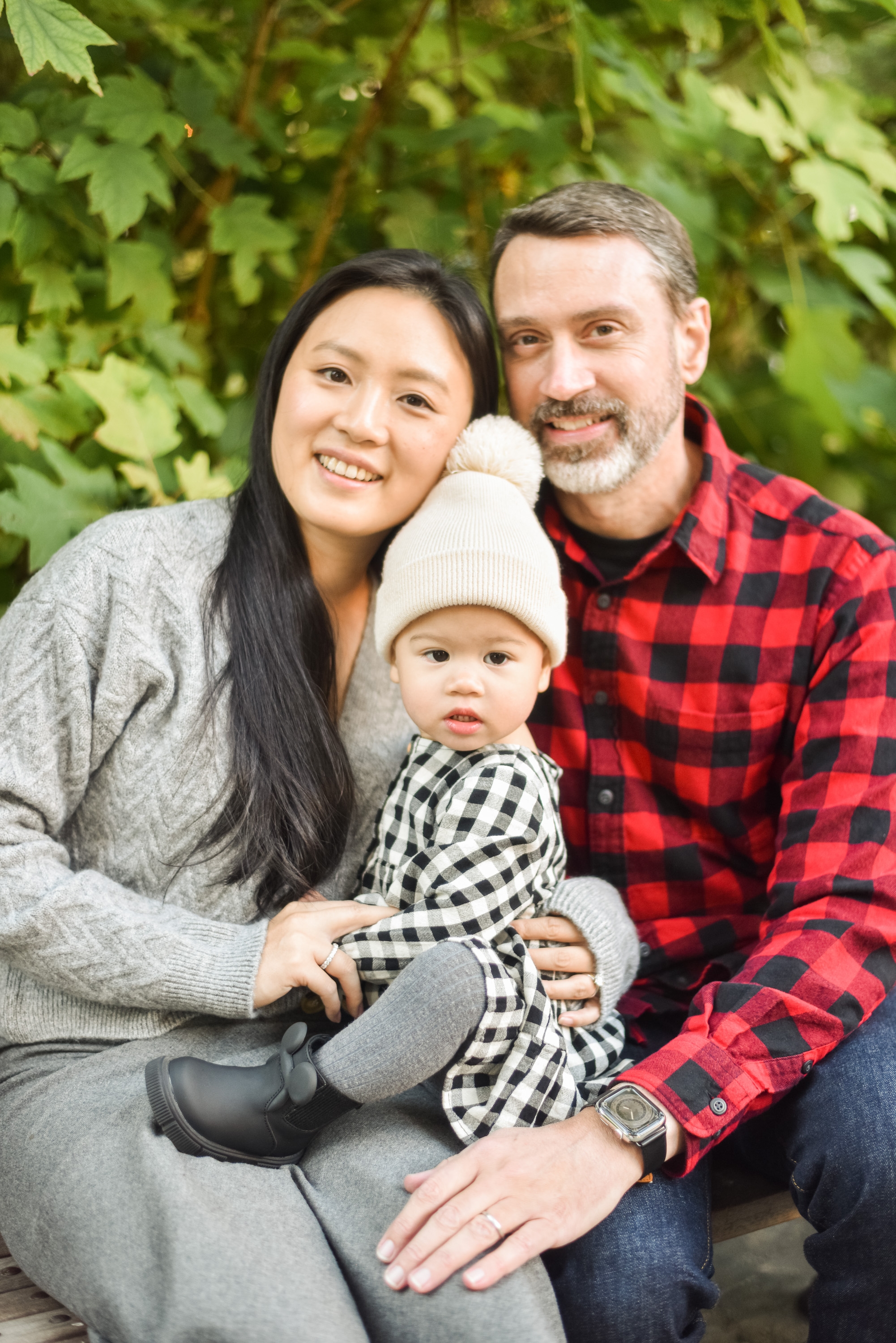 A smiling family of three sits together against lush green foliage, with the mother in gray, father in red plaid shirt, and toddler in white hat and checkered outfit