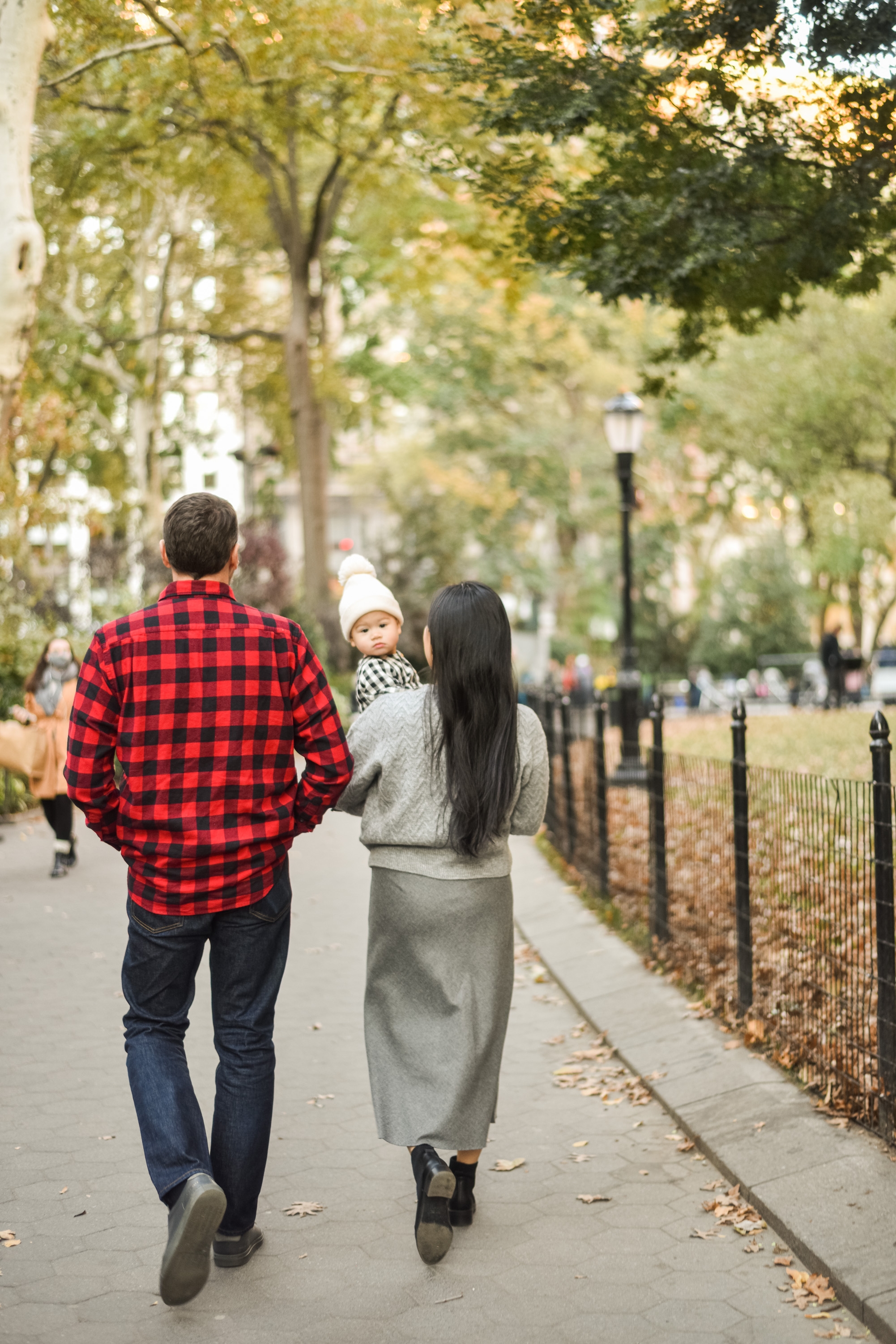 A couple walks with their small child down a tree-lined park path in autumn, the man wearing a red plaid shirt and the woman carrying a child in a white beanie