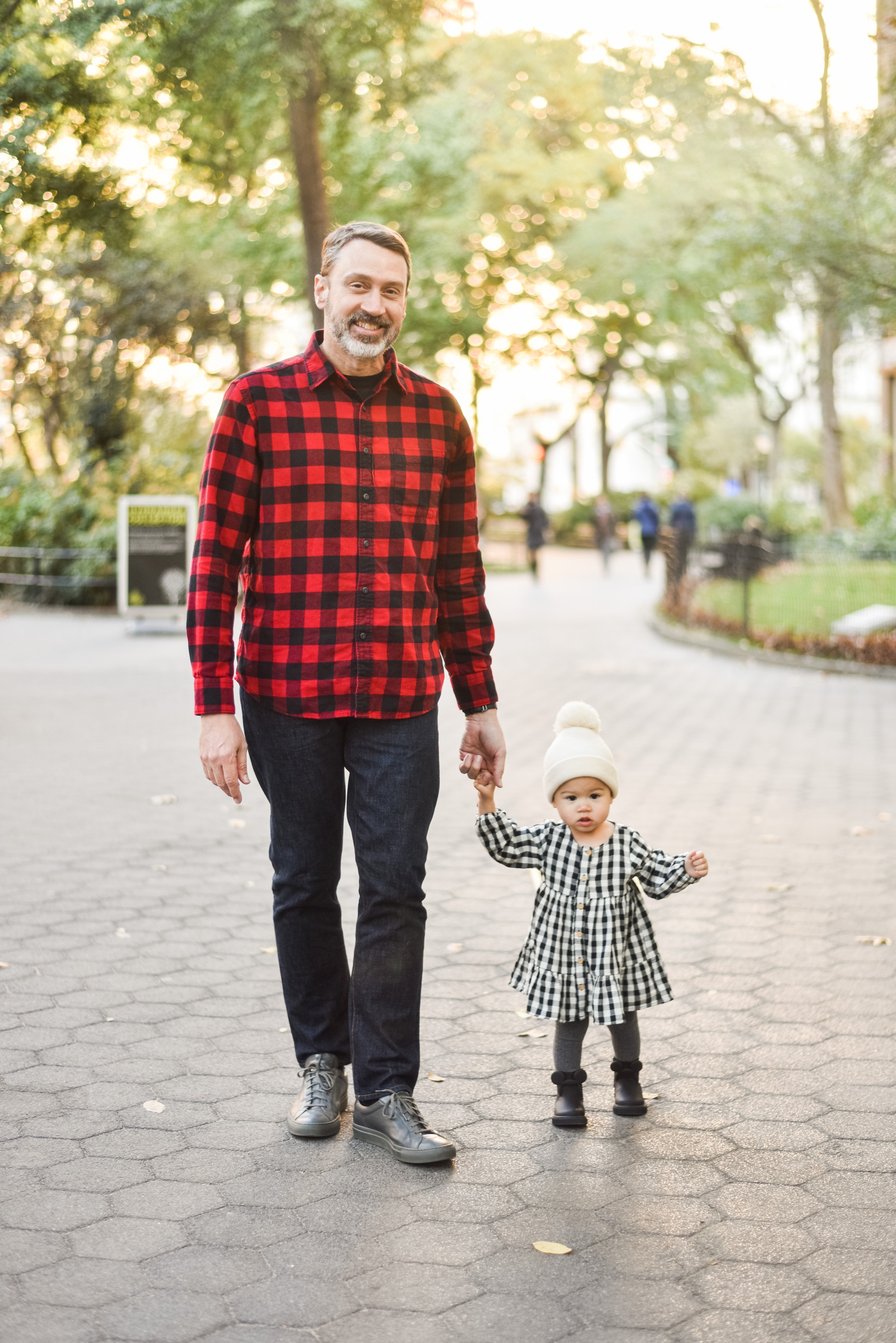 A man wearing a red plaid shirt walks hand-in-hand with a toddler in a white beanie through a tree-lined urban park, bathed in golden afternoon sunlight